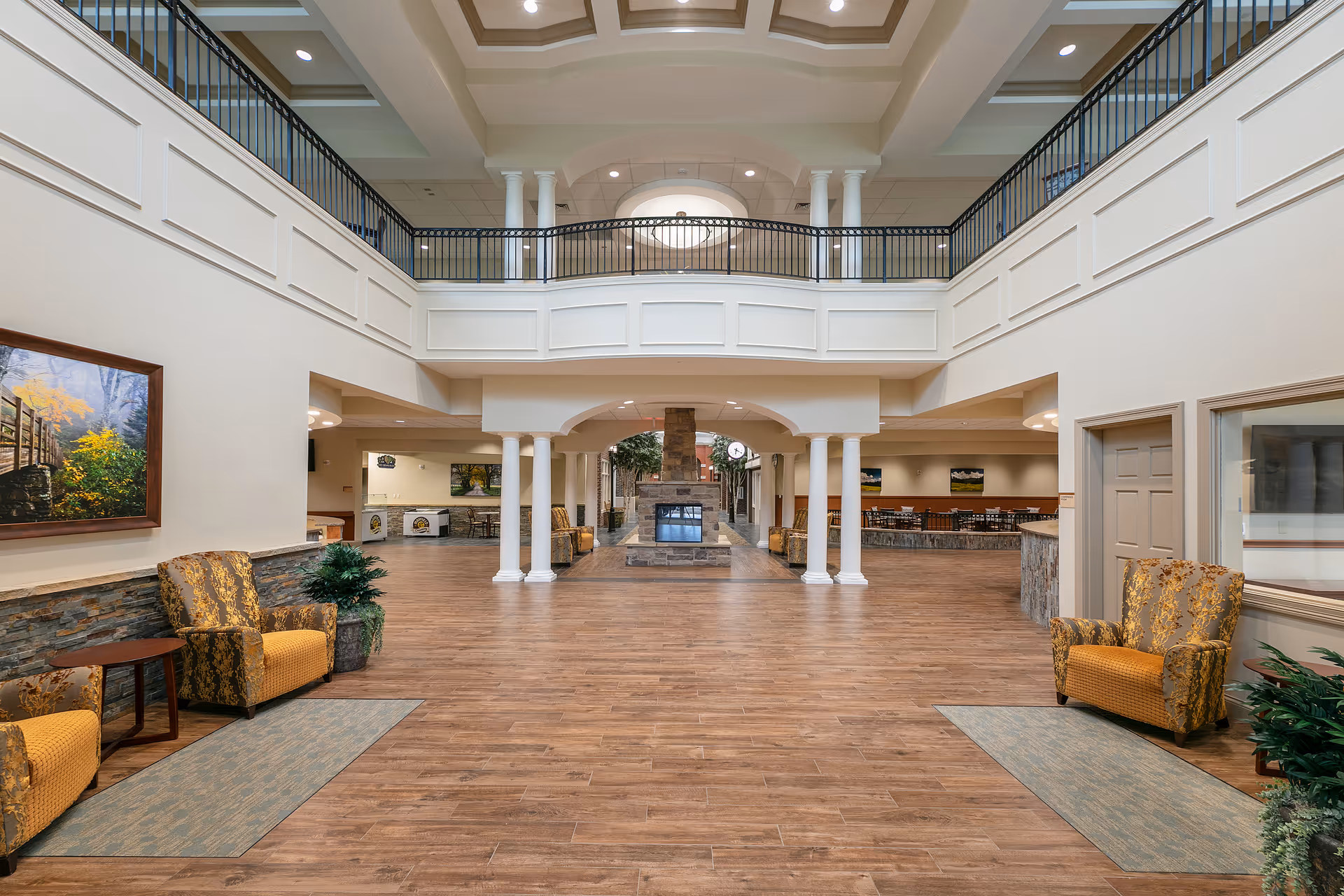 Spacious and well-lit common area in a senior living facility with high ceilings, a second-floor balcony with black railings, and a central stone fireplace. The room features wooden flooring, comfortable patterned armchairs with small side tables, potted plants, and framed artwork on the walls. There are multiple seating areas and a dining area visible in the background.