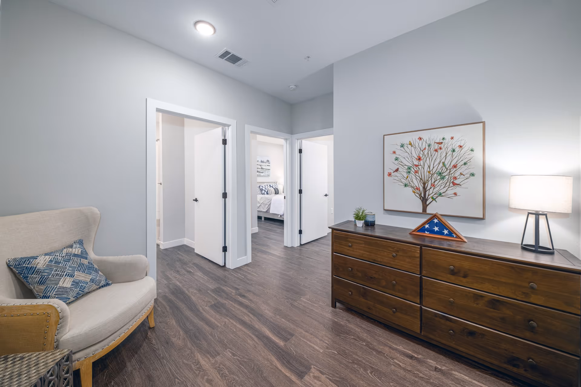 Interior hallway area in a senior living facility with wood flooring, a beige armchair with a blue patterned pillow on the left, and a wooden dresser with a lamp, a small plant, a candle, and a folded American flag in a triangular display case on the right. Three white doors are visible, one leading to a bedroom with a bed and artwork on the wall.