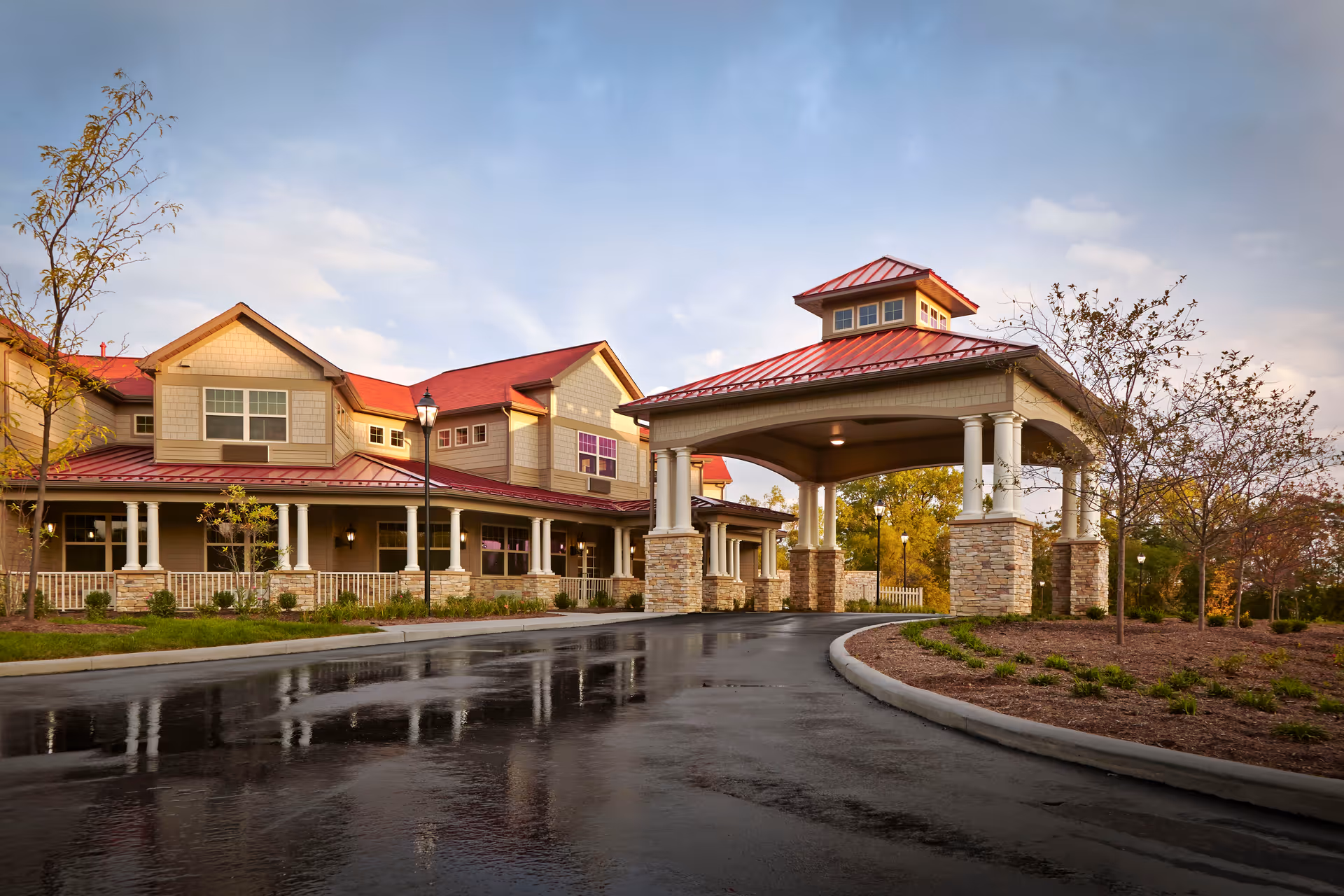 Exterior view of a senior living facility with a covered entrance supported by stone and white columns, a red roof, and a paved driveway surrounded by landscaped greenery and trees under a partly cloudy sky.