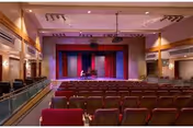 Auditorium with rows of seats facing a stage with red curtains and a person at a piano.