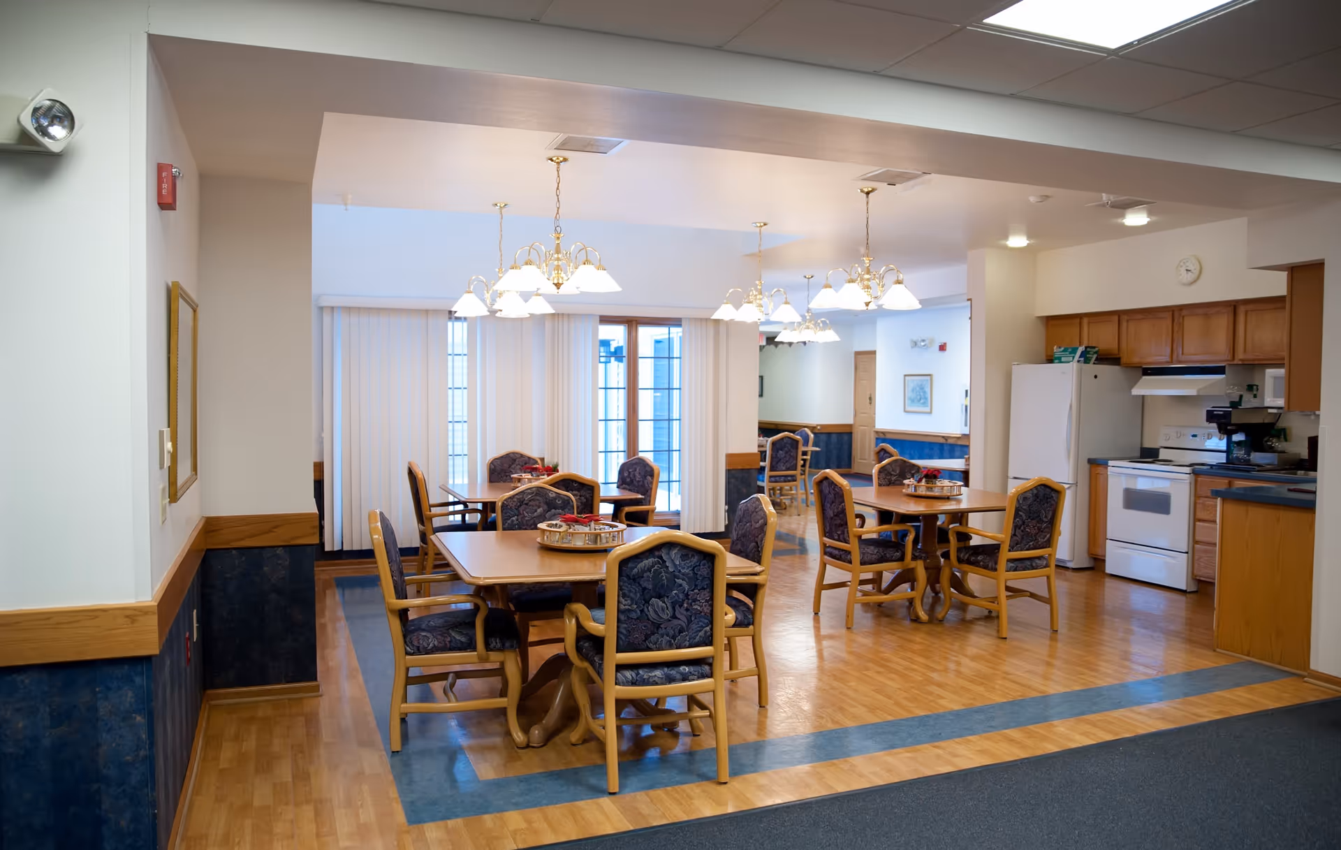 A dining area in Ingleside Manor featuring multiple wooden tables with floral upholstered chairs. The room has hardwood floors with a blue border, several hanging light fixtures, and large windows with vertical blinds. A kitchen area with white appliances and wooden cabinets is visible in the background.