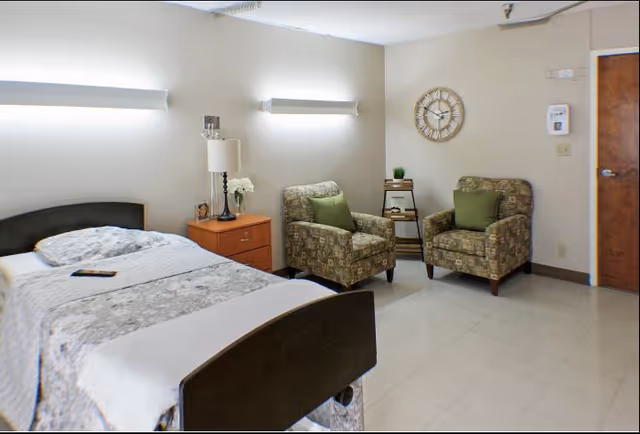 A senior living facility bedroom with a single bed covered in a patterned bedspread, a wooden nightstand with a lamp and flowers, two patterned armchairs with green cushions, a small side table with books and a plant, a wall clock, and a wooden door.