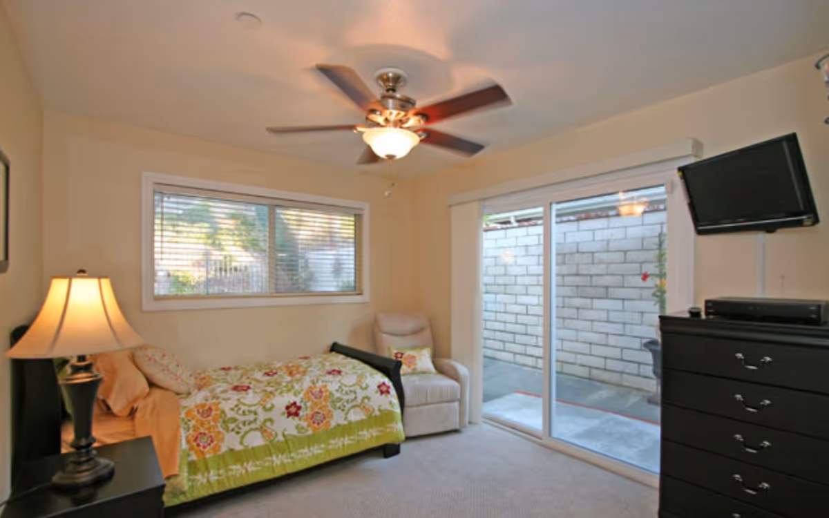 A cozy bedroom with a single bed covered in a floral patterned quilt, a beige armchair with a decorative pillow, a black dresser with a mounted flat-screen TV above it, a table lamp on a nightstand, a ceiling fan with a light, a window with blinds, and sliding glass doors leading to an outdoor patio with a brick wall.
