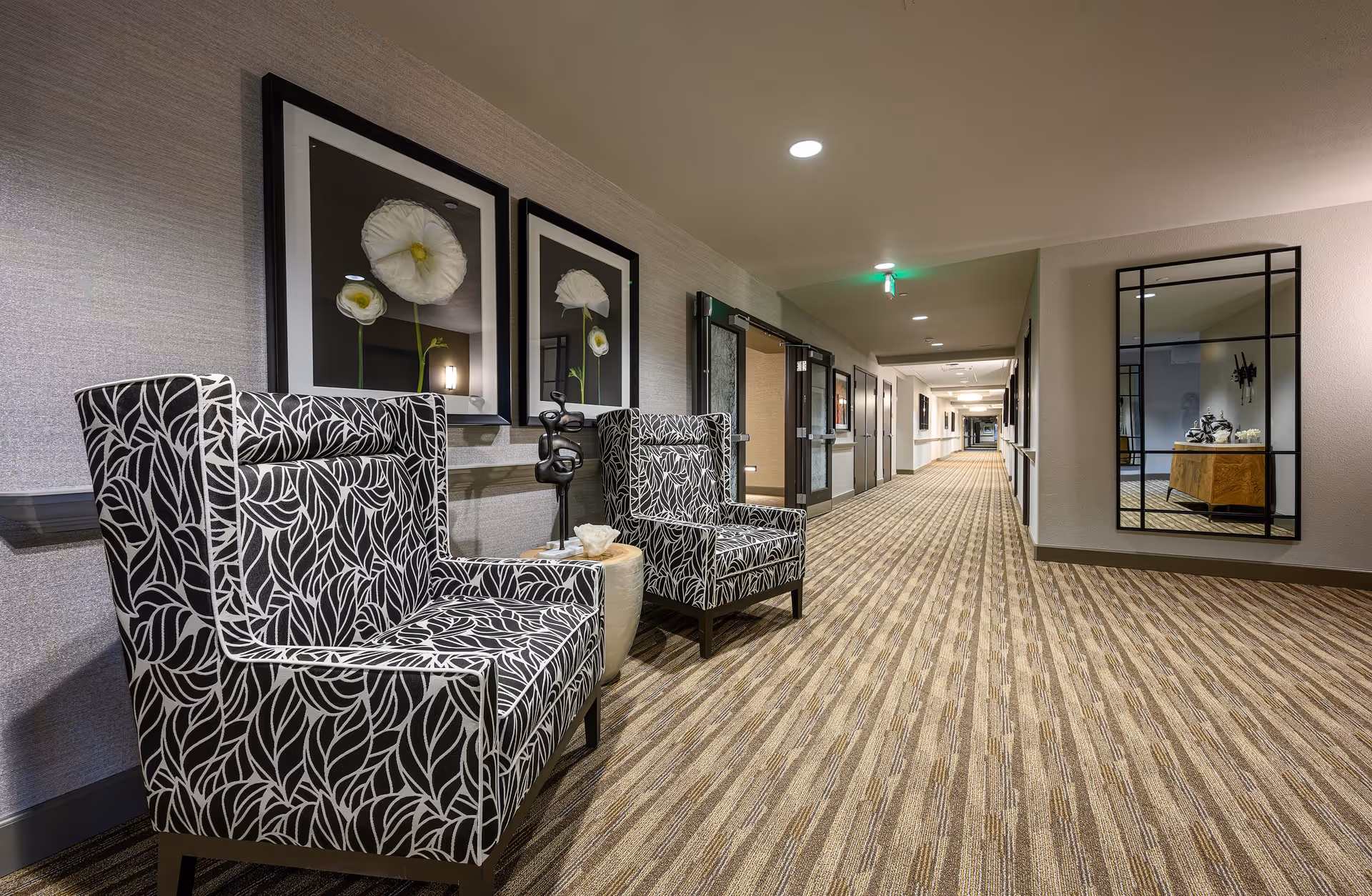Well-lit carpeted interior hallway with patterned armchairs, wall art, and a long corridor of doors.