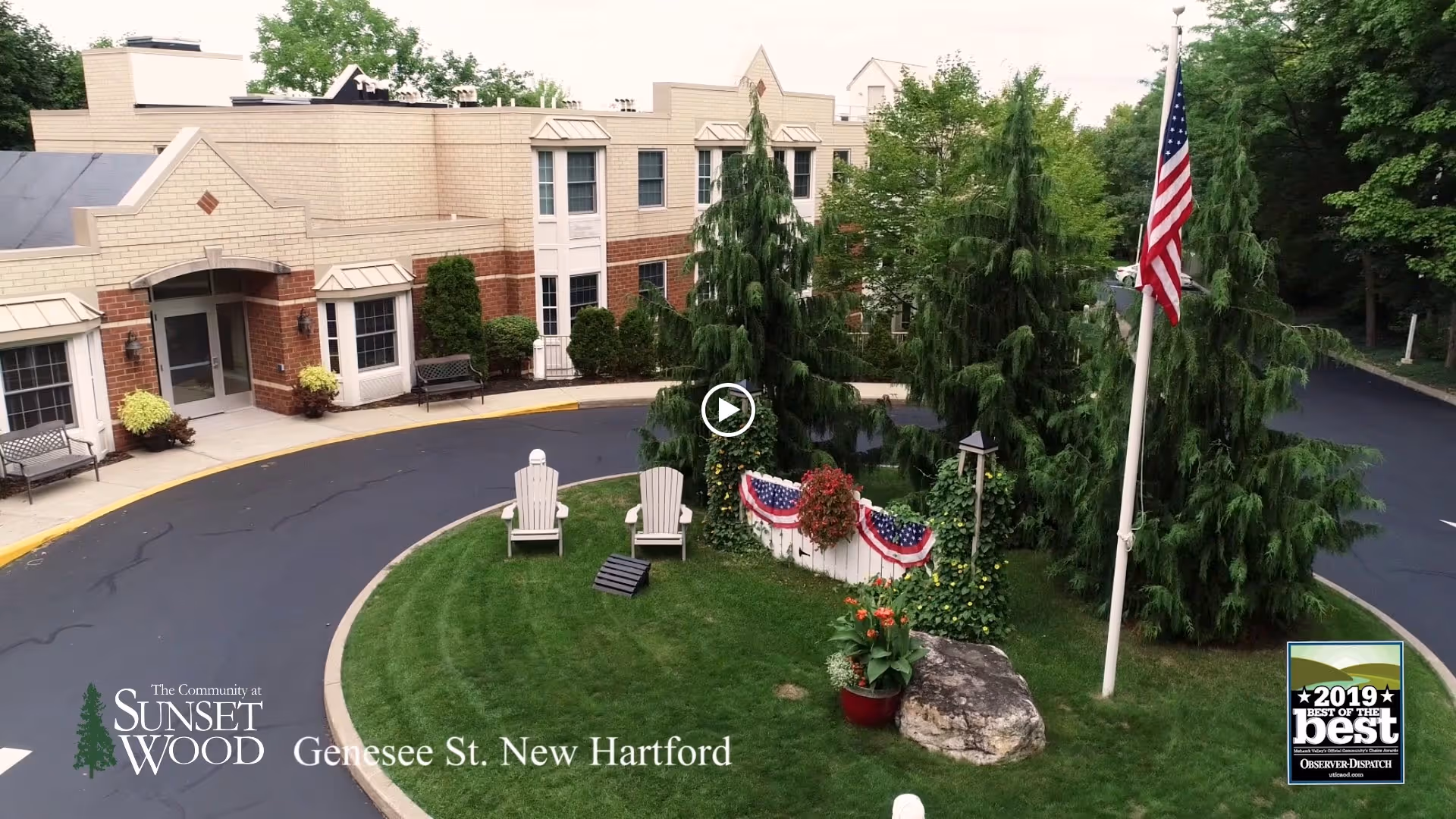 Front entrance of The Community at Sunset Wood with a circular driveway, lawn with Adirondack chairs, landscaping and an American flag in front of the building.