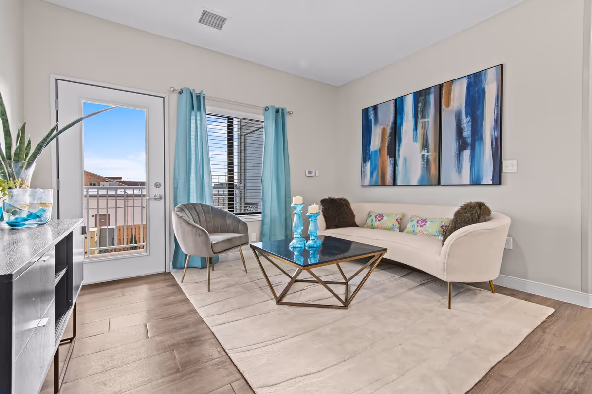 A modern living room with a curved beige sofa adorned with colorful and brown cushions, a gray upholstered chair, a black coffee table with blue candle holders, a large beige area rug, and a sideboard with a plant in a glass vase. The room has light-colored walls, a door with a window leading outside, and a window with turquoise curtains. Three abstract blue and brown paintings hang on the wall above the sofa.
