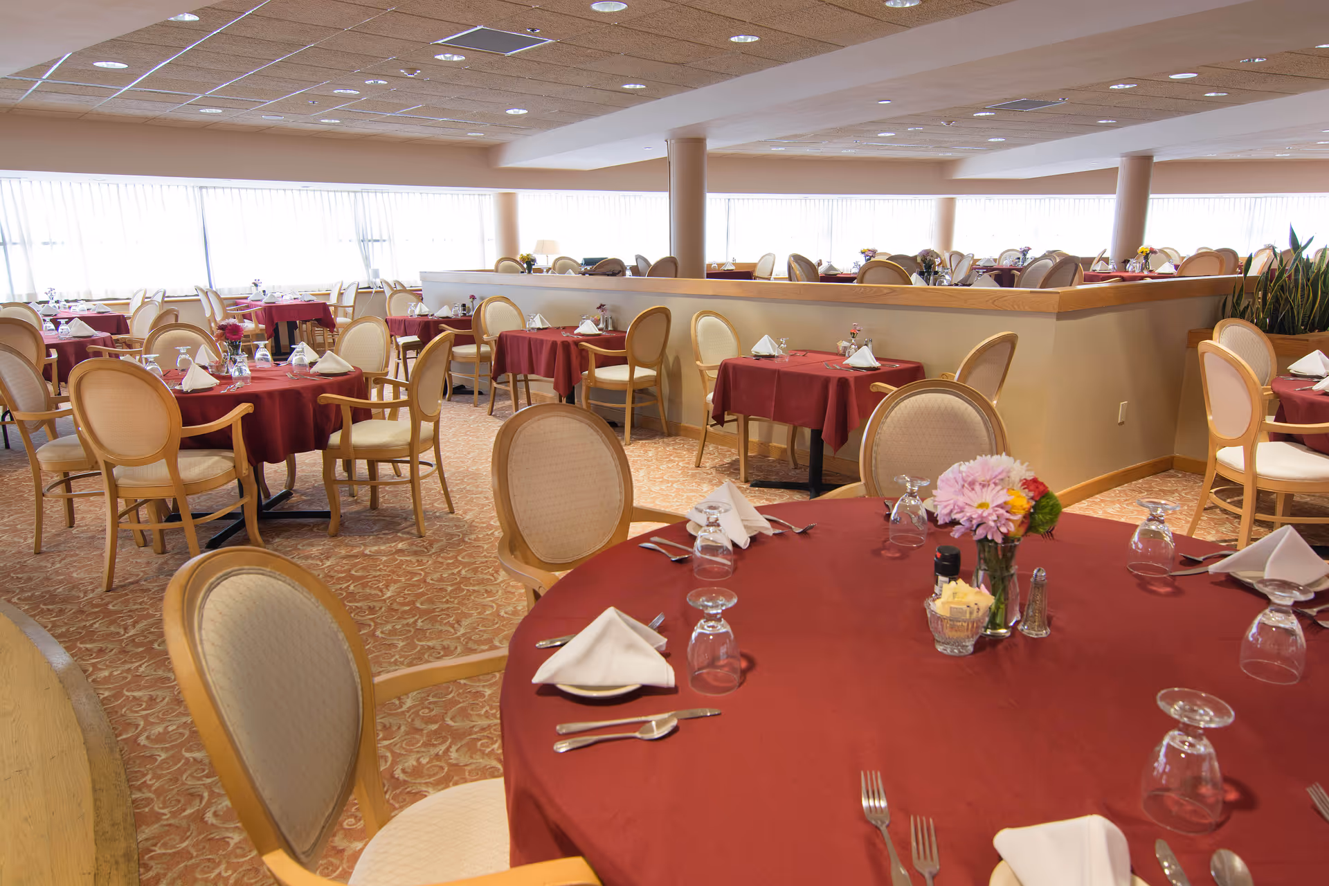Spacious dining room with round tables covered in red tablecloths, set with napkins, glassware, and floral centerpieces.