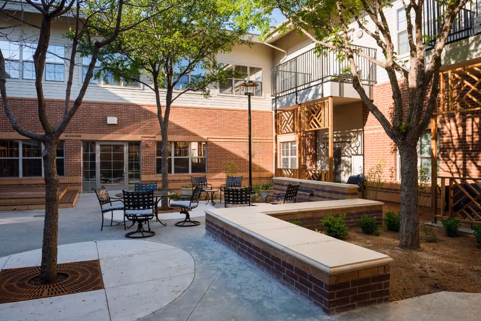 Outdoor courtyard area at The Harrington at Park Central featuring brick walls, several trees, metal chairs and tables, and built-in brick benches with a concrete top. The space is surrounded by windows and doors leading into the building.