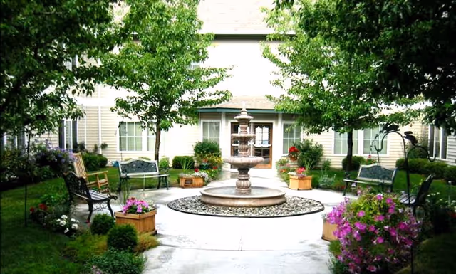 A landscaped courtyard with a central multi-tiered fountain, benches, potted flowers, and trees in front of a building.