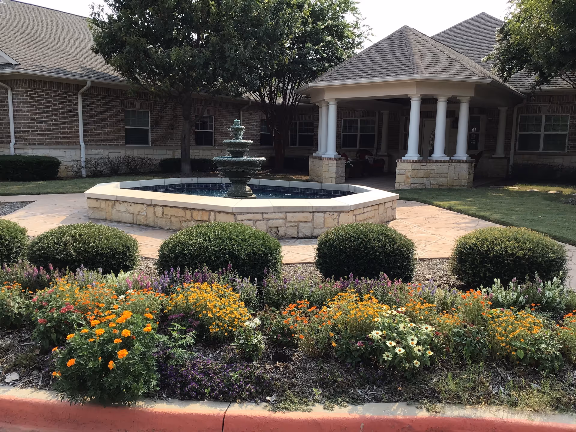 Outdoor garden area at Magnolia Assisted Living and Memory Care featuring a tiered water fountain surrounded by a stone border, neatly trimmed bushes, colorful flower beds, and a covered patio with white columns in the background.