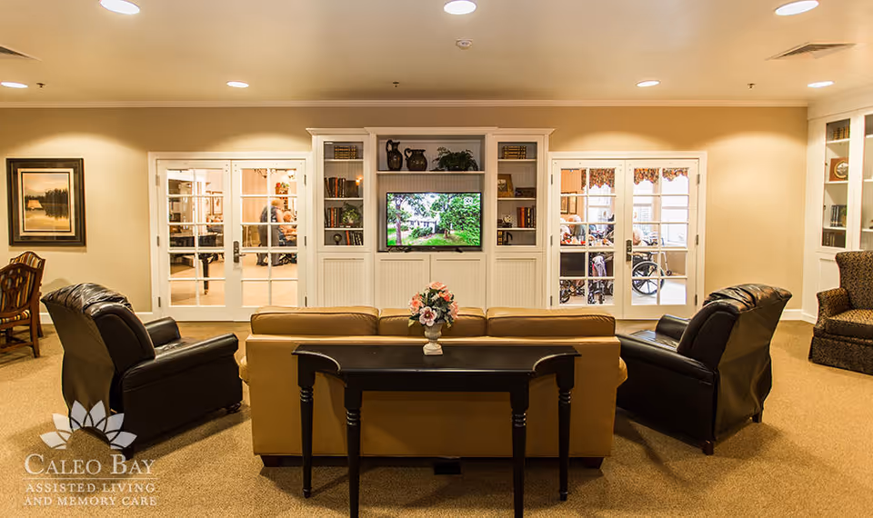 A cozy living room area in an assisted living facility featuring a beige sofa with a small table and flower arrangement behind it, two black leather armchairs, a built-in white entertainment center with shelves holding books and decorative items, and a flat-screen TV displaying a nature scene. French doors in the background reveal other rooms with people, including one person in a wheelchair.