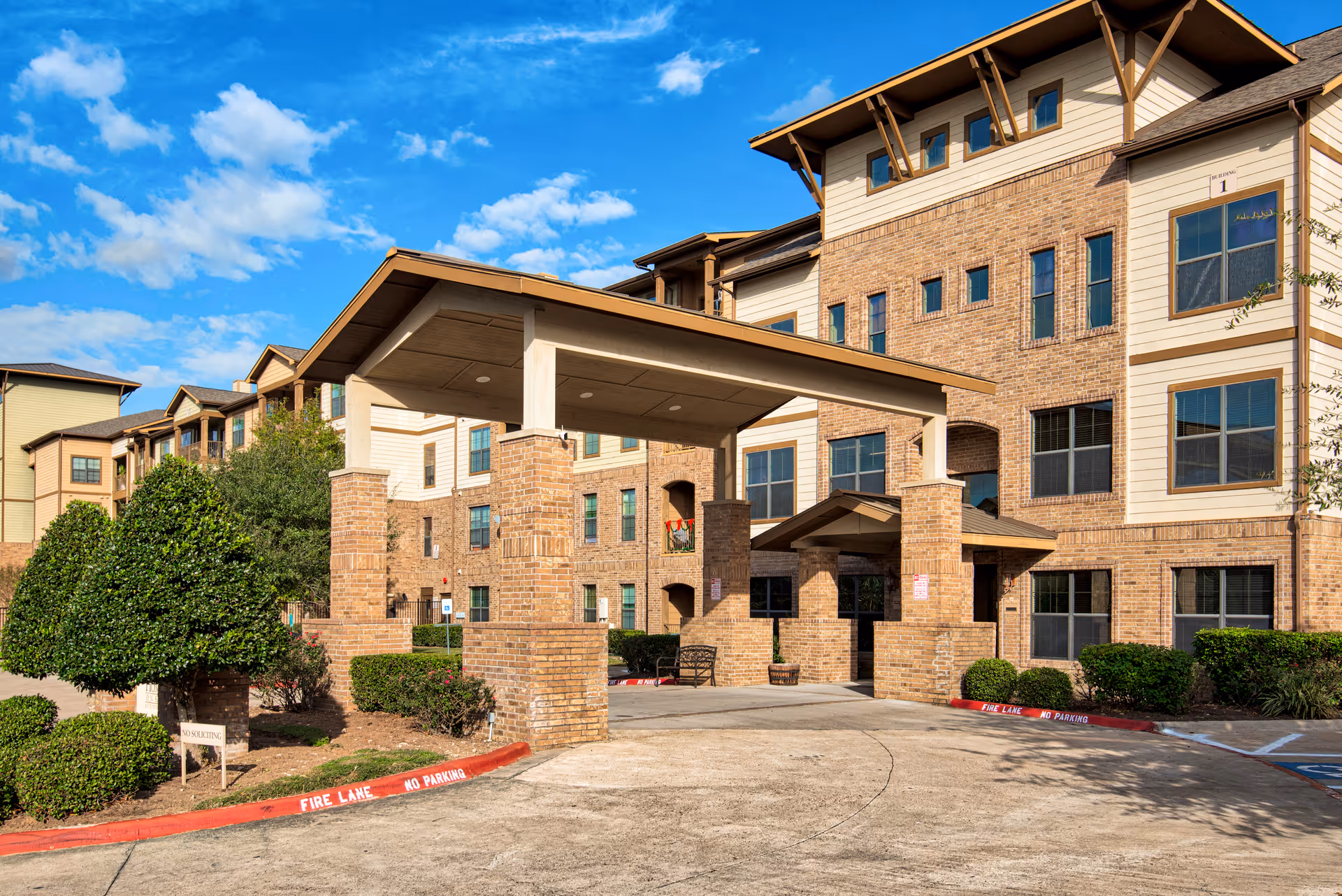 Exterior view of a multi-story senior living facility building with a covered entrance supported by brick pillars, surrounded by neatly trimmed bushes and a clear blue sky above.