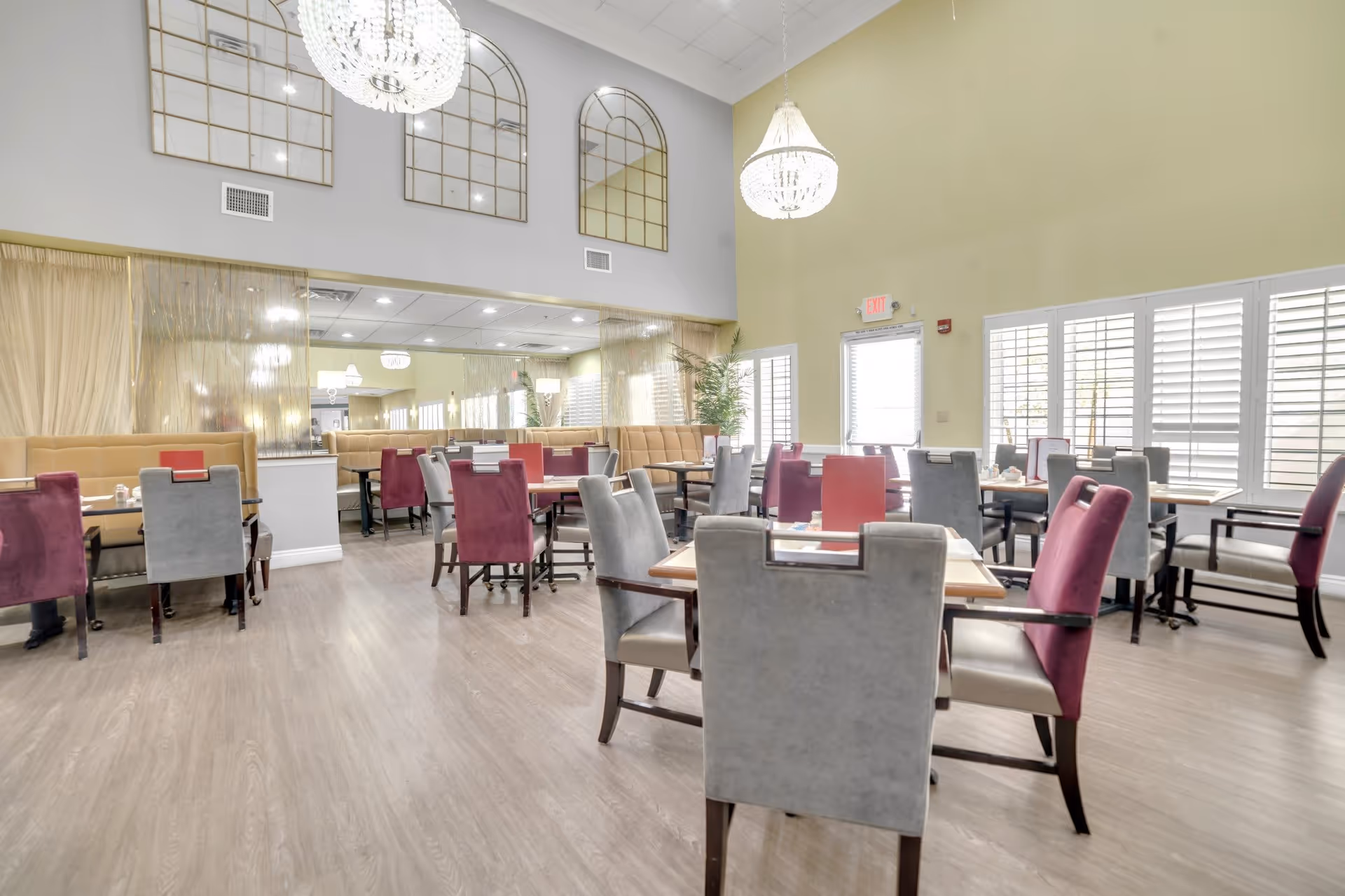 Bright dining room with multiple tables and chairs in shades of gray and purple, beige cushioned bench seating along the walls, large windows with white shutters, two elegant chandeliers hanging from a high ceiling, and decorative wall mirrors.