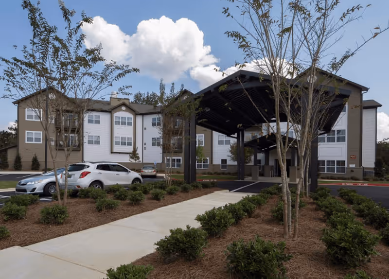 Exterior view of a three-story senior living facility with a covered entrance driveway, landscaped bushes, young trees, and parked cars under a partly cloudy sky.