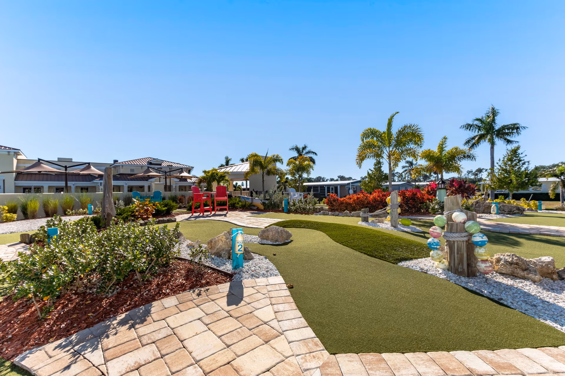 Outdoor miniature golf course with artificial turf, surrounded by landscaping including bushes, palm trees, and colorful plants. There are red chairs and umbrellas in the background under a clear blue sky.