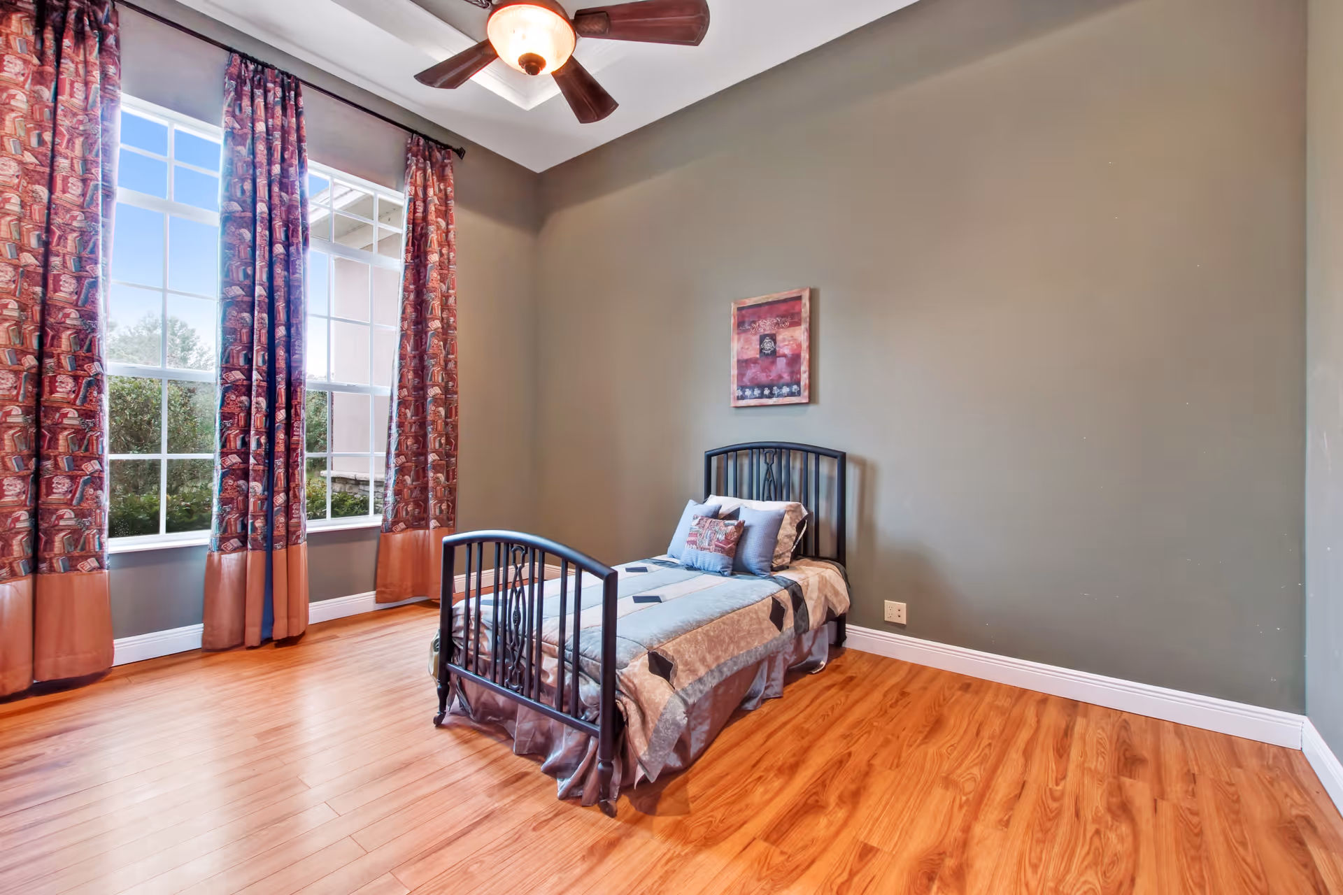 A simple bedroom with a single metal bed frame, a neatly made bed with pillows, wooden flooring, large windows with patterned curtains, a ceiling fan with light, and a framed picture on the wall.
