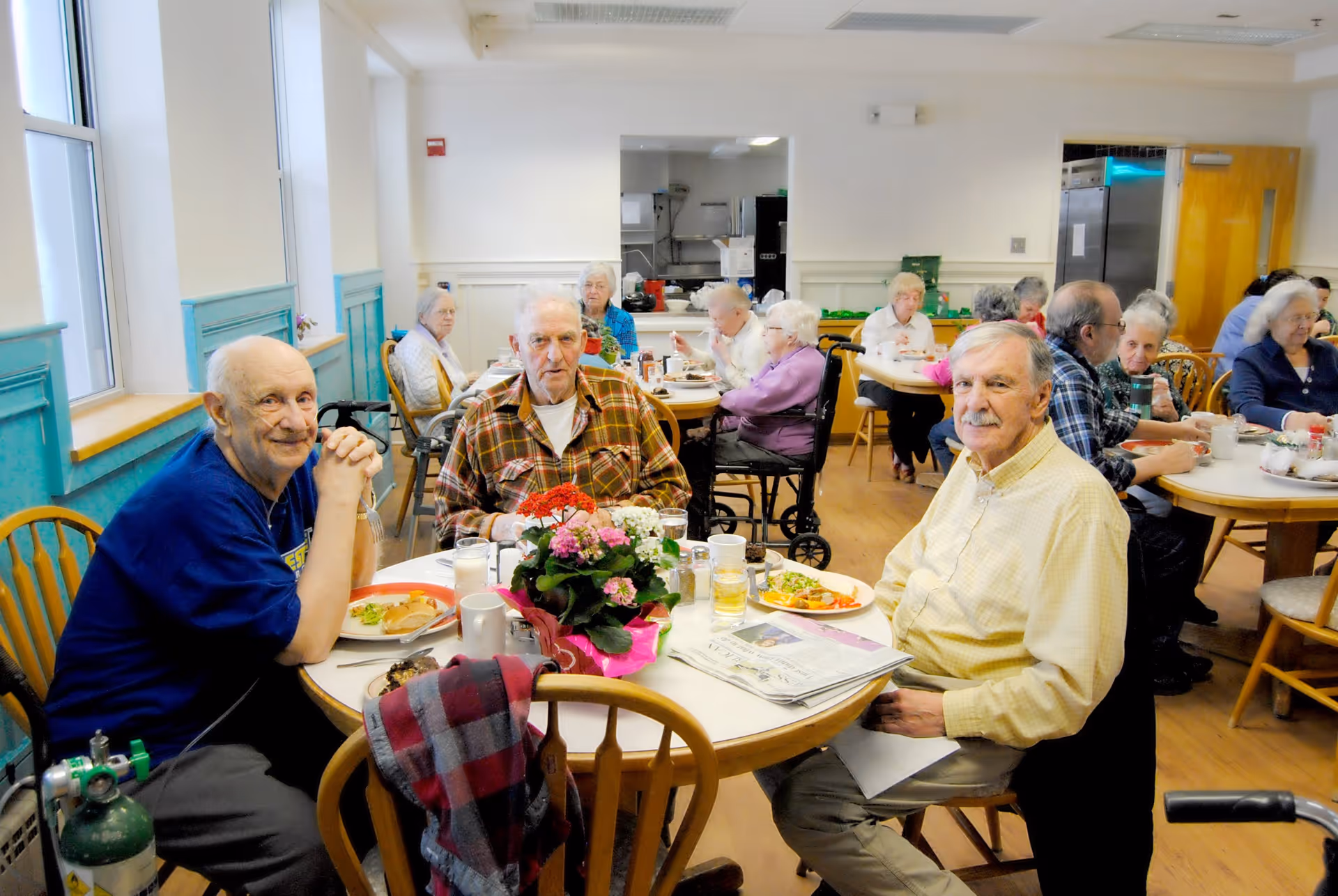 A group of elderly people sitting around tables in a dining room at Keene Valley Neighborhood House, eating meals and socializing. The room has light-colored walls with teal wainscoting, wooden chairs, and large windows letting in natural light. There are flowers on the tables and some individuals using mobility aids.