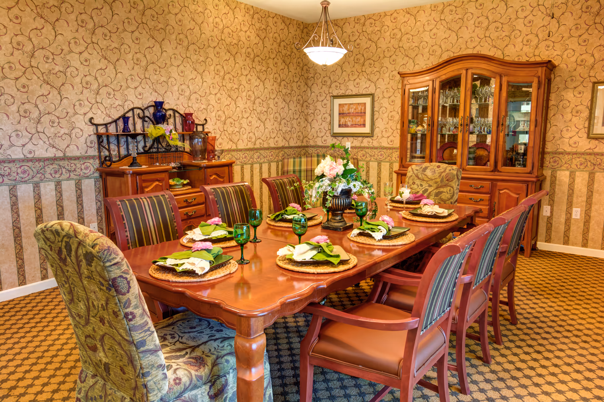 A formal dining room with a long wooden table set for eight people. The table is adorned with green napkins, pink napkin rings, woven placemats, and green glassware. There is a floral centerpiece in the middle of the table. The room features patterned wallpaper with a floral and striped design, a wooden china cabinet filled with glassware, and a sideboard with decorative items. The carpet has a geometric pattern, and the lighting is provided by a ceiling fixture.
