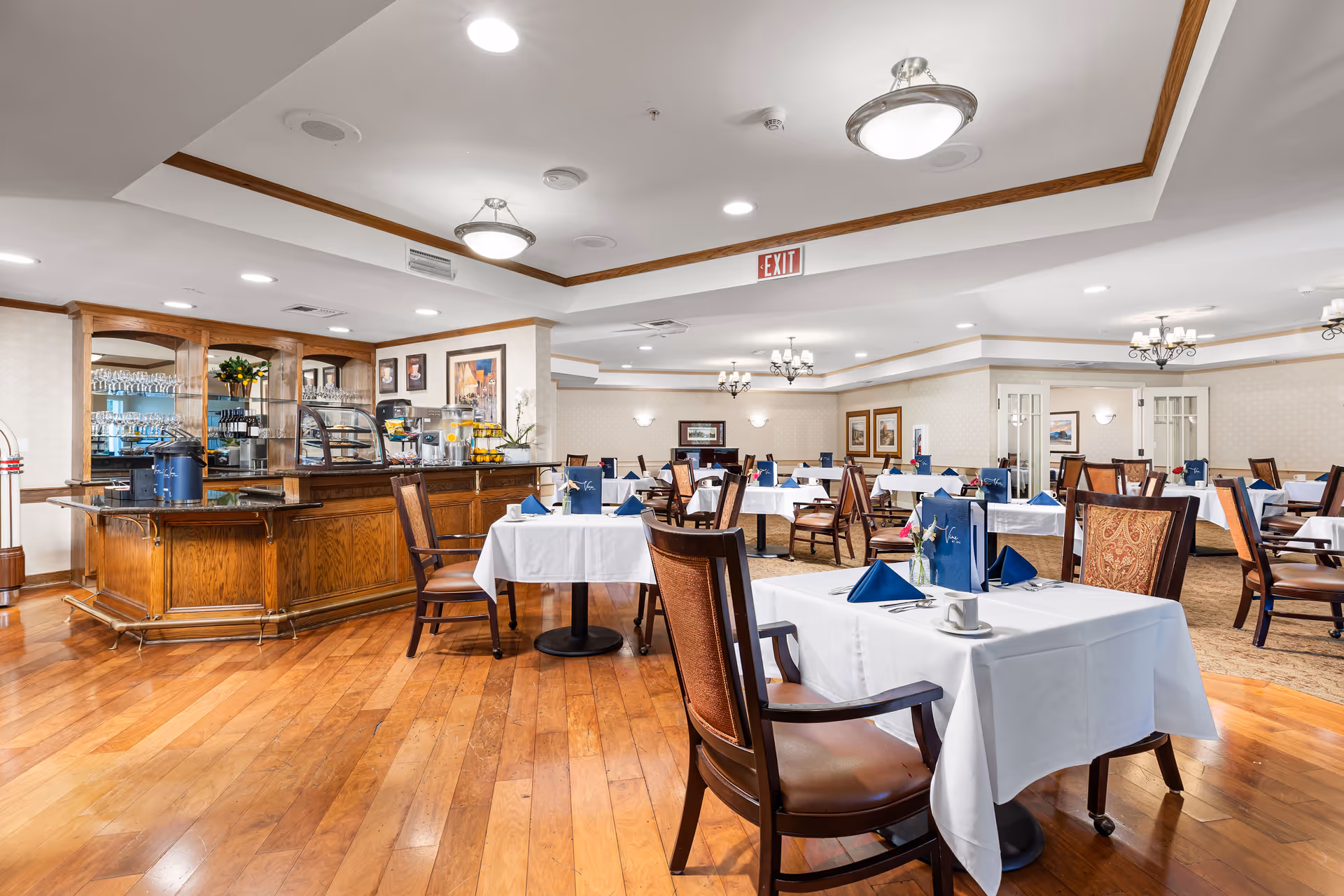 Spacious dining room with wood floors, neatly set tables with white linens and blue napkins, and a wooden service counter.