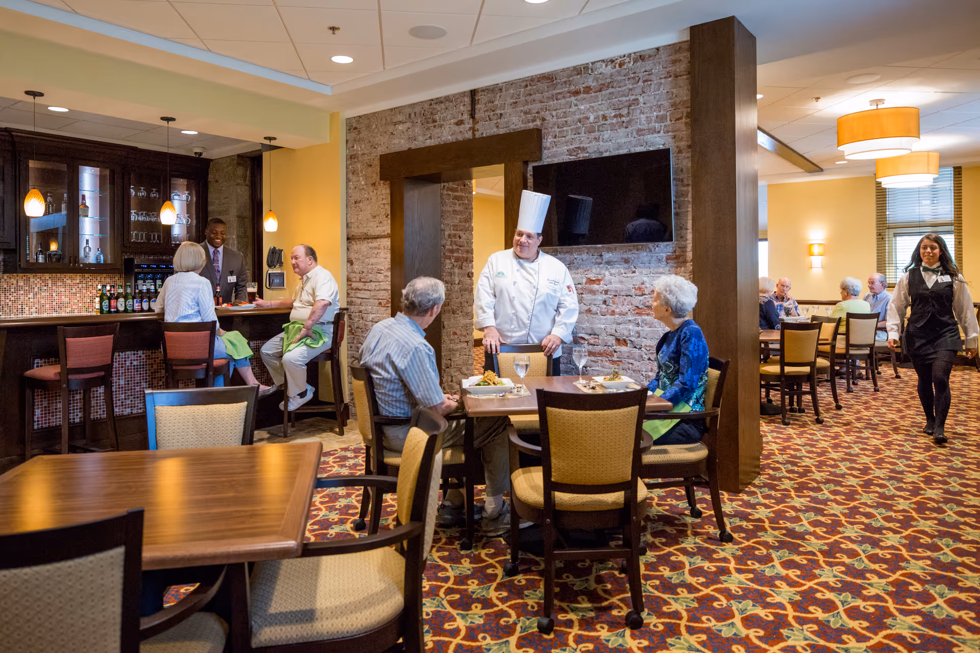 A dining area in a senior living facility with elderly residents seated at tables and a bar. A chef in a white uniform and hat is standing and talking to two seated residents. A server is walking through the room. The room has warm lighting, patterned carpet, and a brick accent wall with a mounted TV.