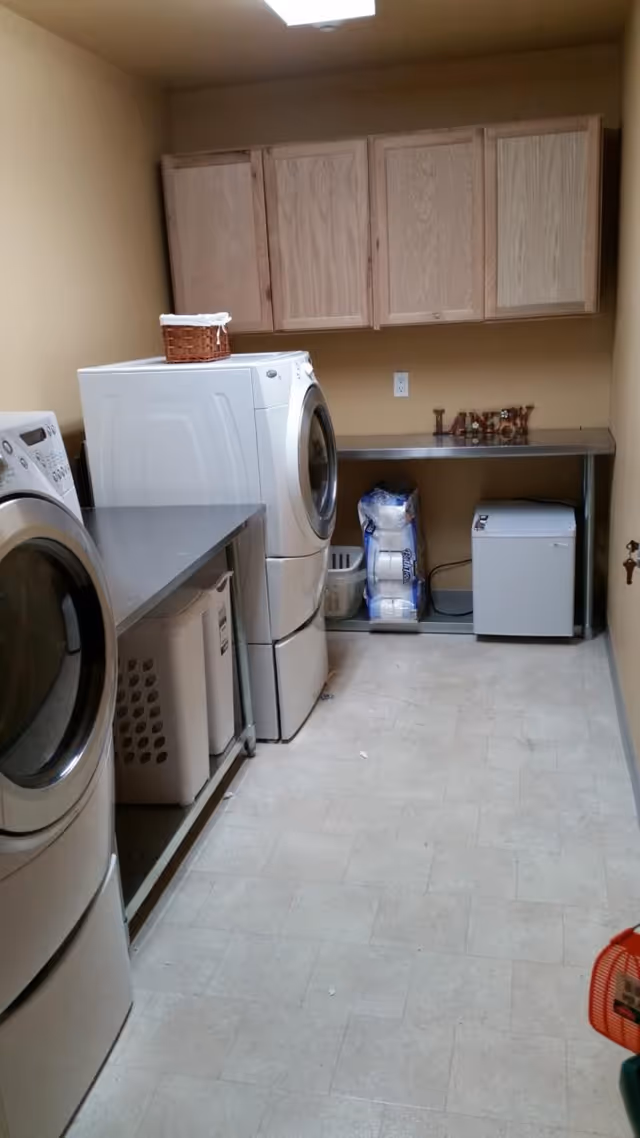 Laundry room with a front-loading washing machine and dryer, a metal countertop with laundry baskets underneath, wooden cabinets mounted on the wall, a small freezer, and a pack of paper towels on the floor.