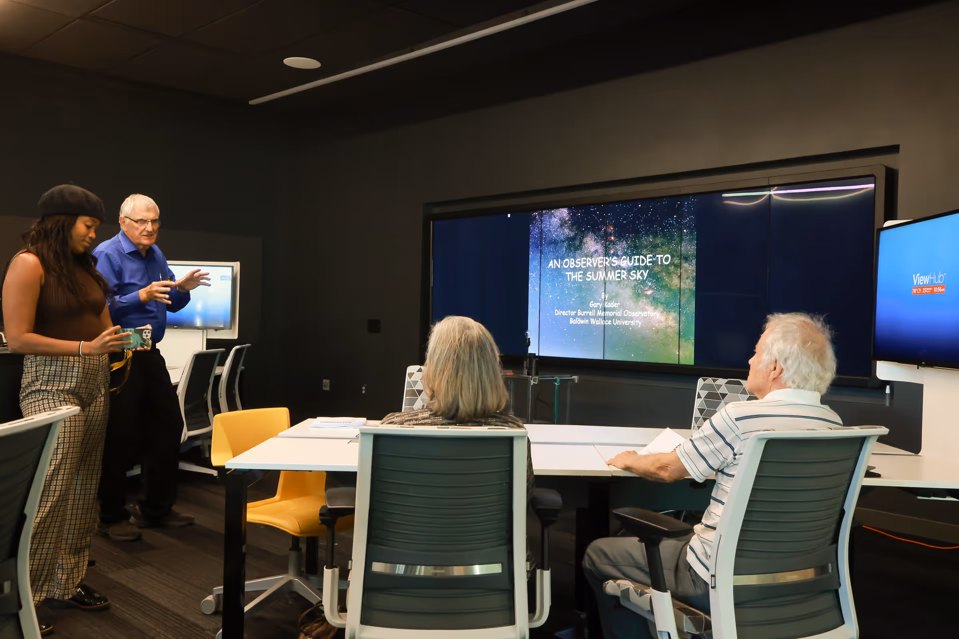 A group of four people in a modern conference room. Two elderly individuals are seated at a table facing a large screen displaying a presentation titled 'An Observer's Guide to the Summer Sky.' A man in a blue shirt is standing and speaking, while a younger woman stands nearby holding a cup. The room has office chairs and a dark ceiling.