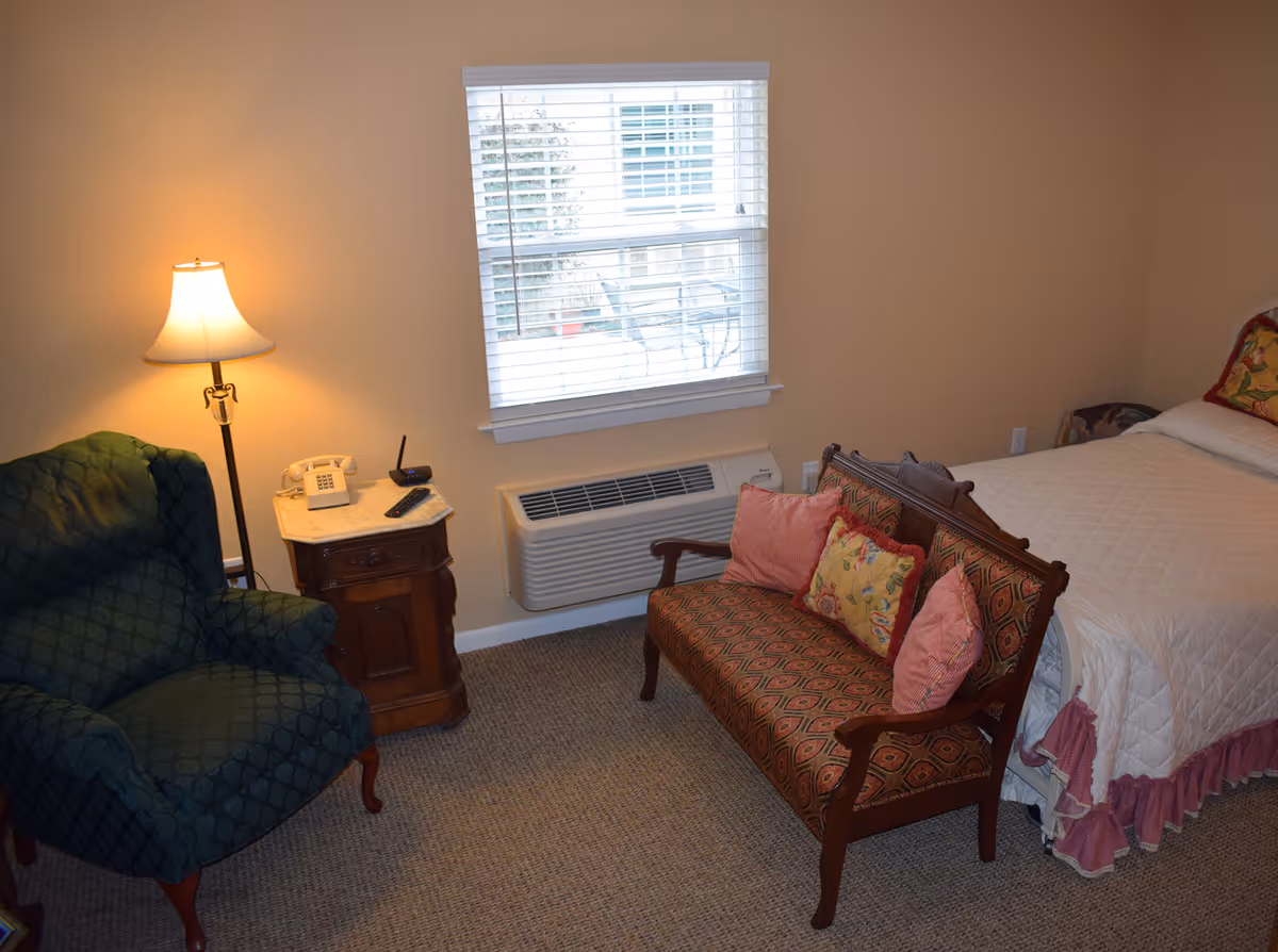 Furnished bedroom with a bed, patterned settee and armchair, a nightstand with lamp and telephone, and a window-mounted HVAC unit.