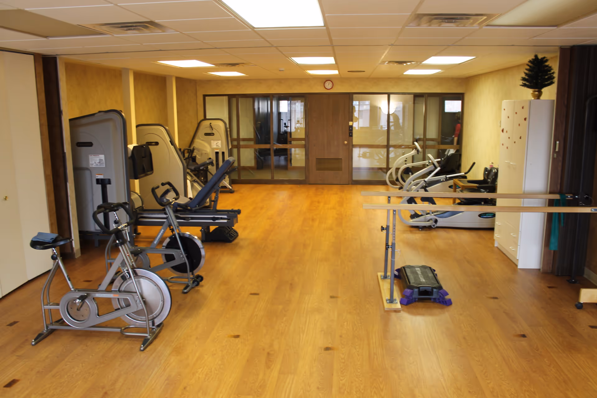 Exercise room with stationary bikes, fitness machines and parallel bars on a wood floor in a senior living facility.