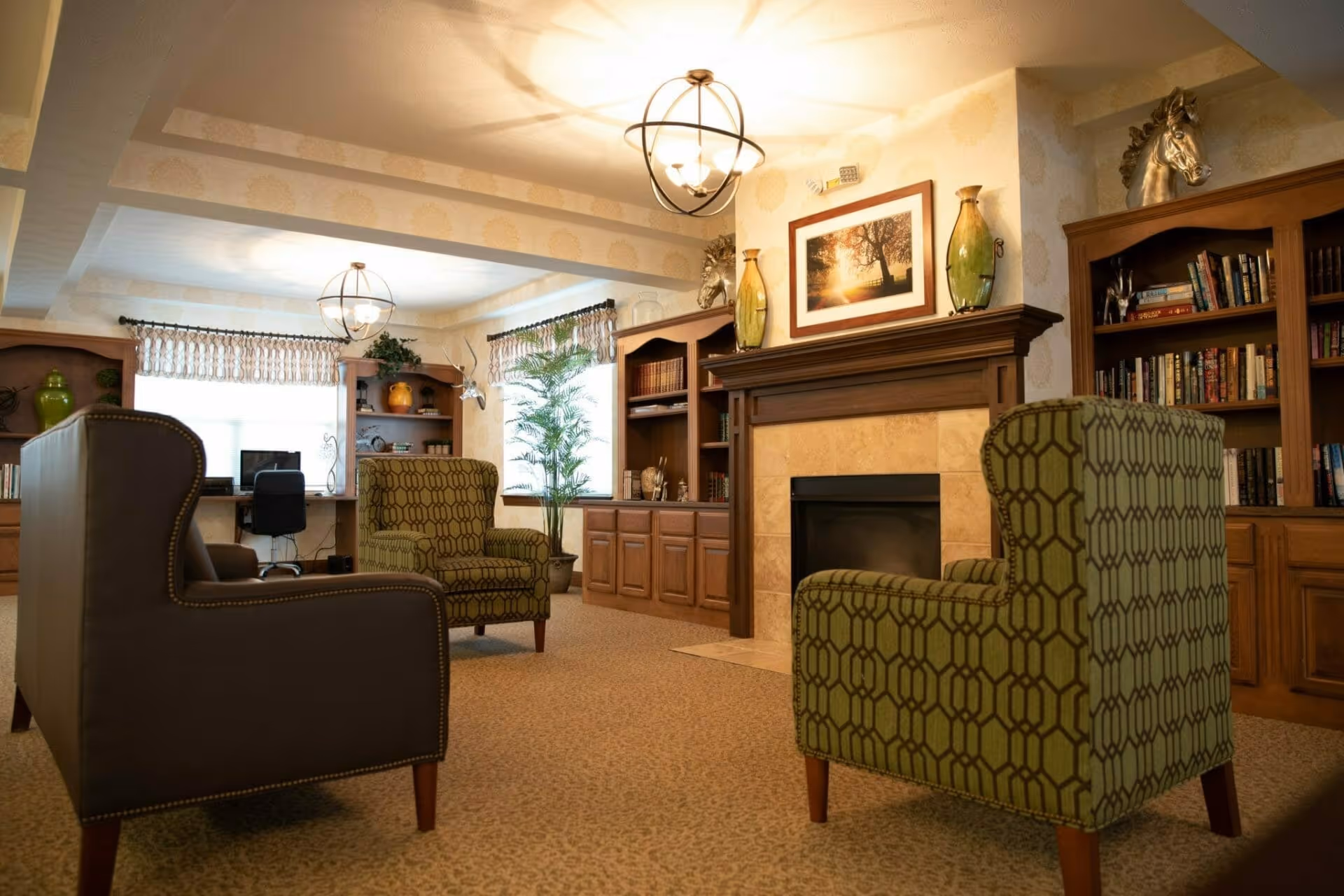 A cozy living room area with two green patterned armchairs and one dark brown armchair arranged around a fireplace with a wooden mantel. The mantel is decorated with two large vases and a framed picture of a tree at sunset. Built-in wooden bookshelves filled with books and decorative items flank the fireplace. In the background, there is a desk with a computer and a black office chair near two windows with patterned valances. The room is warmly lit by ceiling light fixtures and has a beige carpeted floor.