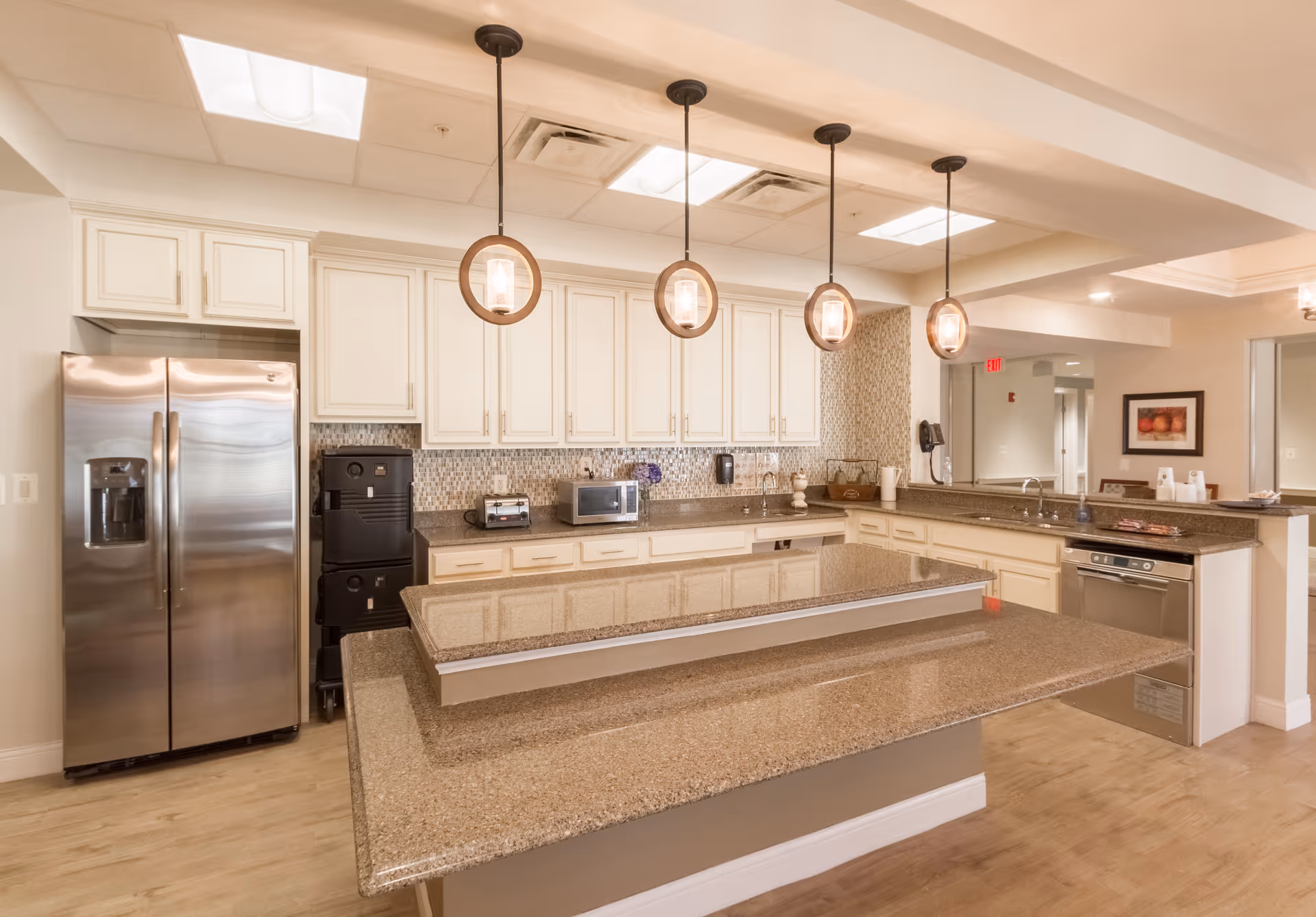 A modern kitchen area with beige cabinets, a large granite countertop island, stainless steel refrigerator, dishwasher, microwave, toaster, and four pendant lights hanging from the ceiling. The kitchen has a light wood floor and a mosaic tile backsplash.