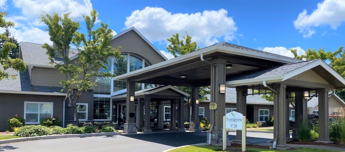 Exterior view of Evergreen Memory Care facility showing a large covered entrance with pillars, a driveway, landscaped greenery, and a clear blue sky with some clouds.
