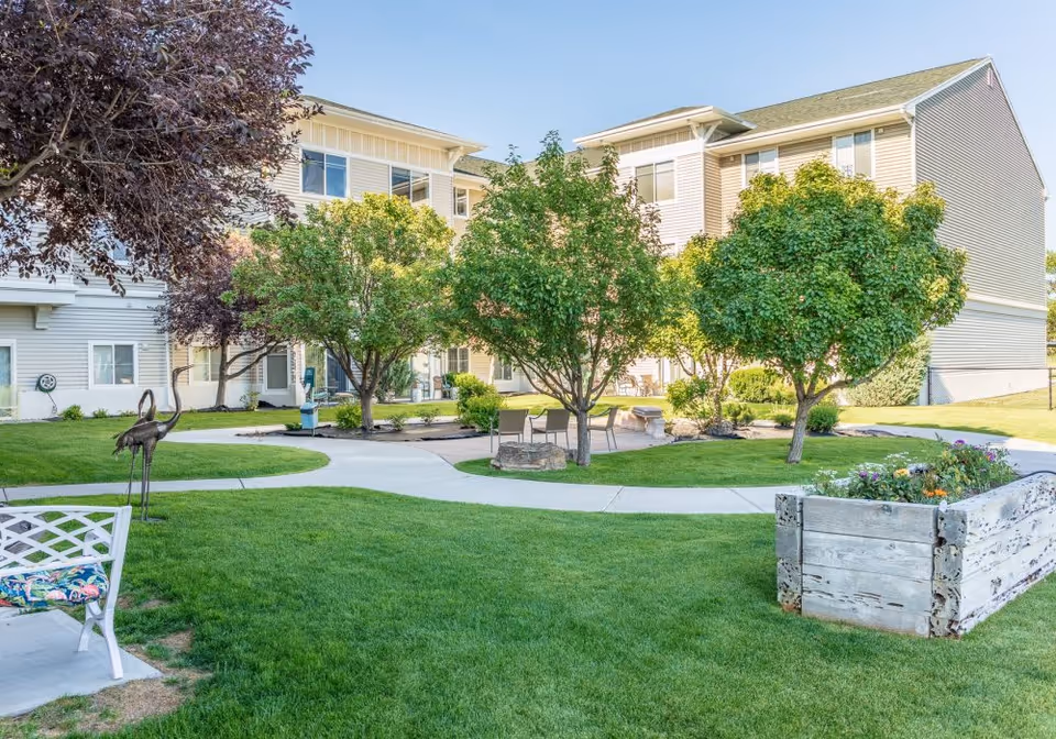 A well-maintained outdoor garden area at a senior living facility with green grass, several trees, a wooden raised flower bed with blooming flowers, a white bench with a colorful cushion, metal bird sculptures, and a paved walkway. The background shows a two-story beige building with multiple windows and a clear blue sky.