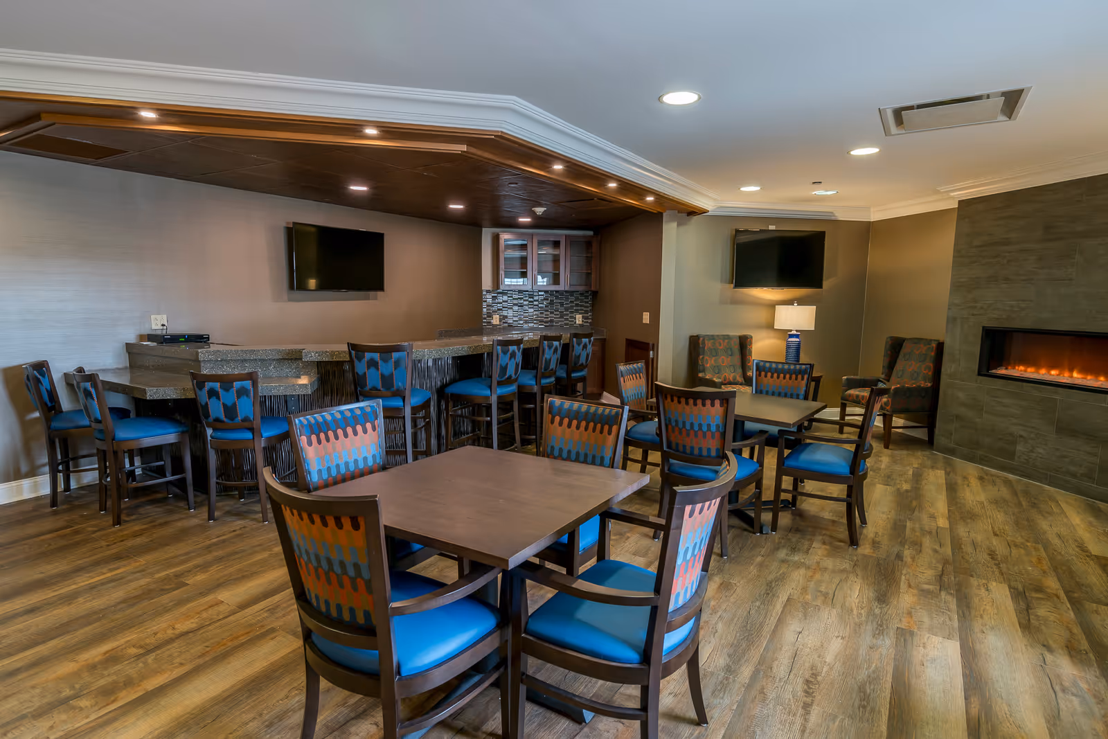 A senior living common room with multiple tables and blue-upholstered chairs, a small bar, wall-mounted TVs, and an electric fireplace.