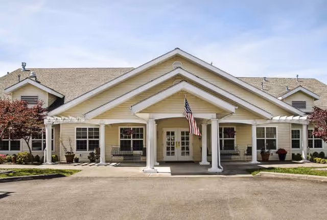 Front exterior view of a single-story assisted living facility building with a covered entrance supported by white columns, an American flag hanging in the center, and landscaped areas with small trees and potted plants on either side.