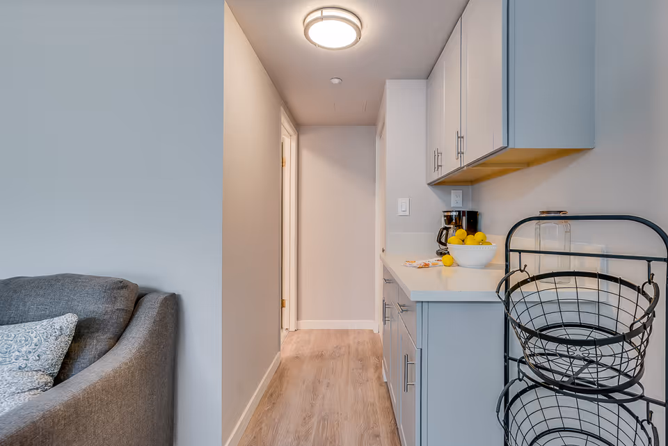 A narrow hallway with light wood flooring leads to a doorway. On the right side, there is a white countertop with cabinets above and below, a coffee maker, a bowl of lemons, and a black metal basket stand. On the left side, part of a gray upholstered sofa with a patterned pillow is visible. The walls are painted light gray, and a round ceiling light fixture illuminates the space.