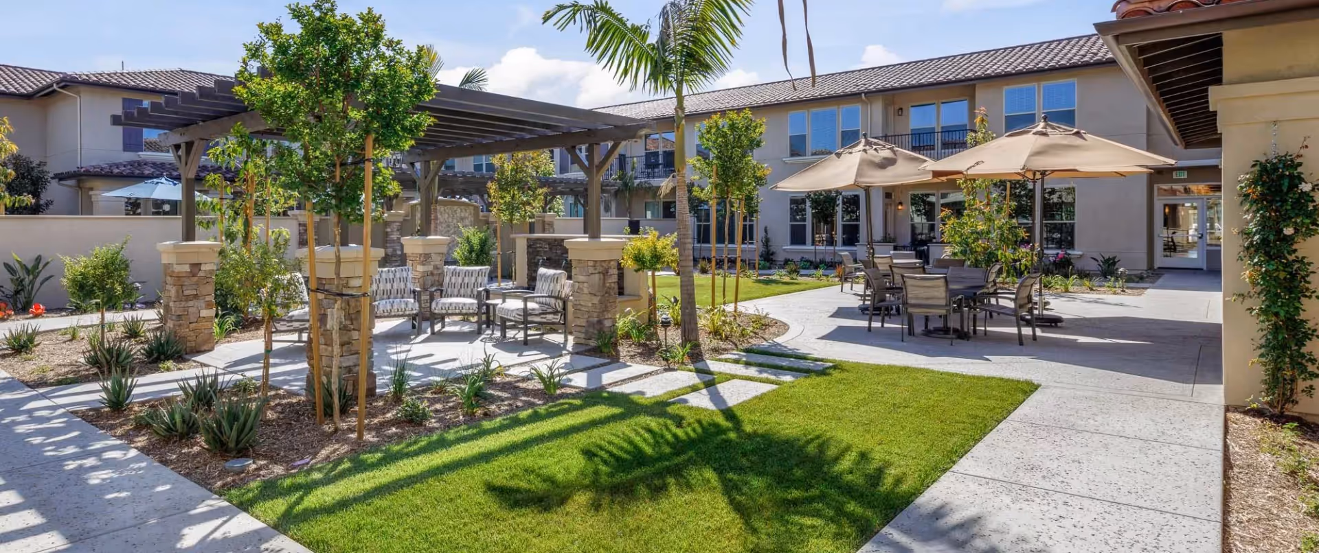 Outdoor courtyard area at Oakmont of Riverpark featuring a pergola with cushioned chairs, several small trees and plants, a grassy lawn, and tables with umbrellas for shade. The building with multiple windows surrounds the courtyard under a clear blue sky.