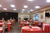 A spacious dining room with multiple round tables covered in red tablecloths, each surrounded by beige chairs. The room is decorated with Christmas decorations including a Christmas tree near the back wall. There is a black stage area on the left side and a television mounted on the right wall. Large windows and glass doors are visible at the back of the room.