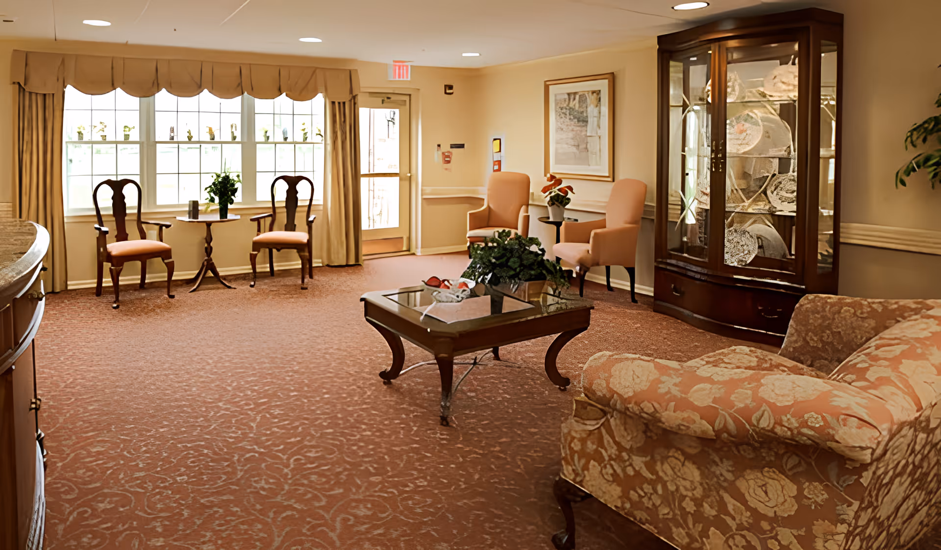 A cozy living room area with patterned carpet, floral upholstered armchair, wooden coffee table with a plant and decorative items, two peach-colored armchairs with a small side table and plant, two wooden chairs with cushions near a window with beige curtains, and a wooden display cabinet with glass doors containing decorative plates.