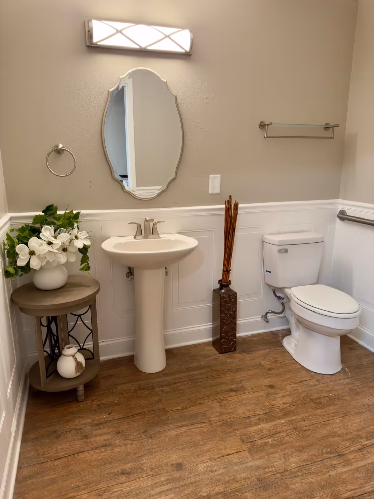 A clean bathroom with a white pedestal sink, an oval mirror above it, a toilet to the right, and a small round wooden table with white flowers in a vase to the left. The floor is wood-style, and the walls are painted beige with white wainscoting. There is a modern light fixture above the mirror.