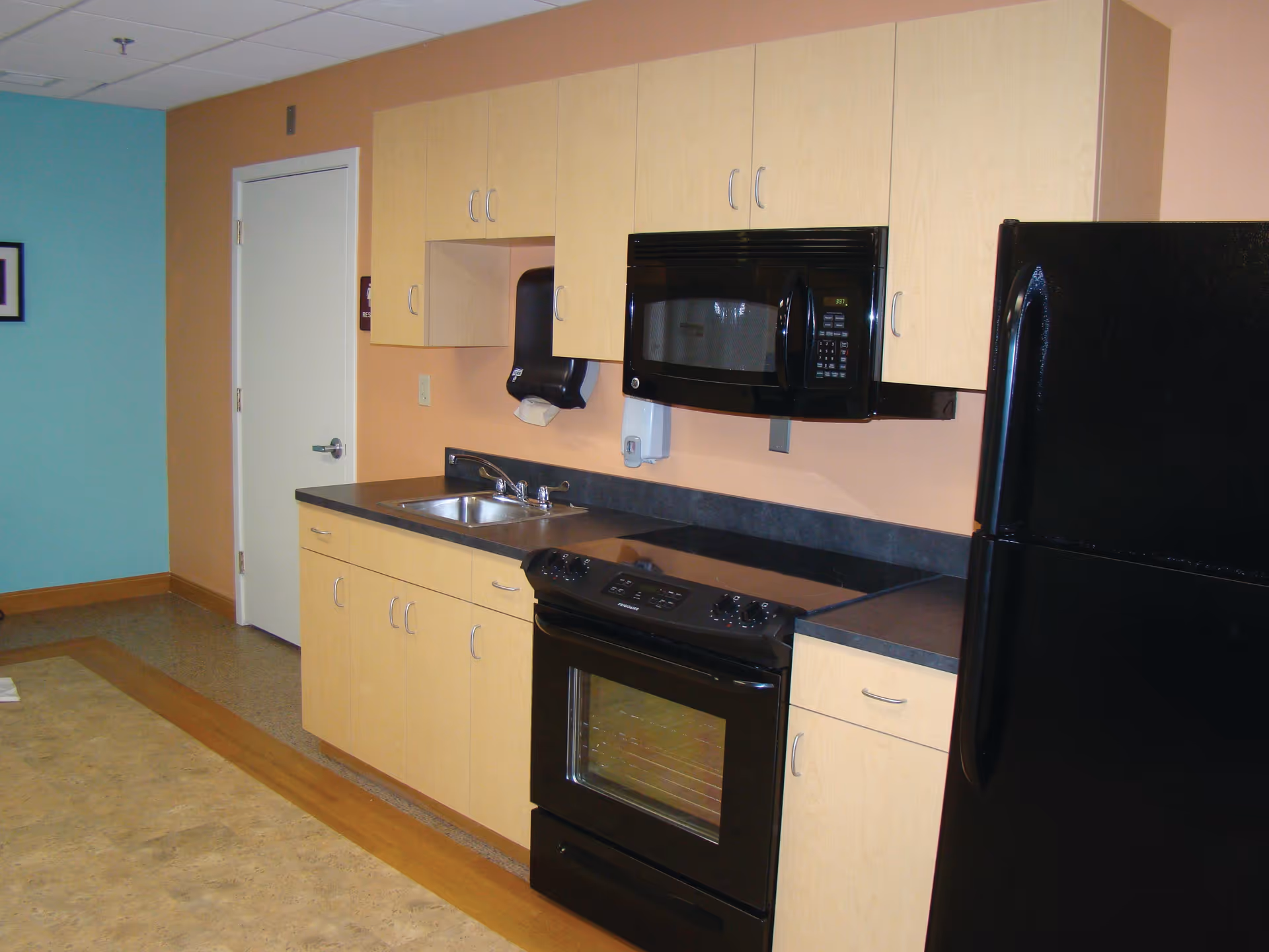 A kitchen area with light wood cabinets, a black microwave mounted above a black stove, a black refrigerator, and a stainless steel sink. The walls are painted in two colors: peach and teal. There is a white door on the left side of the kitchen.