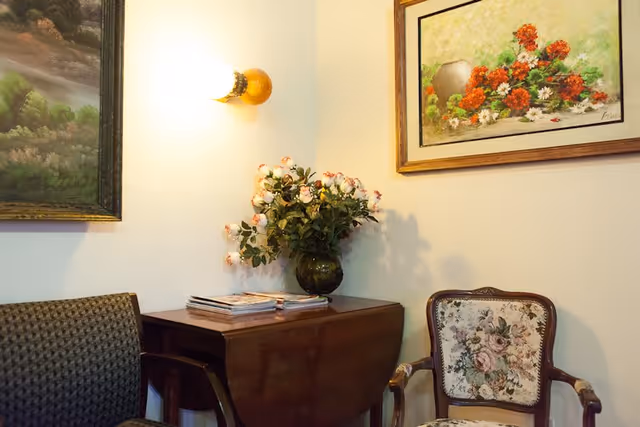 A cozy corner of a room with a wooden drop-leaf table holding a vase of pink and white flowers and some magazines. There are two chairs, one with a floral upholstery and wooden arms, and the other with a dark patterned fabric. The walls are decorated with two framed paintings, one of a landscape and the other of a floral arrangement. A wall-mounted light fixture is turned on above the table.