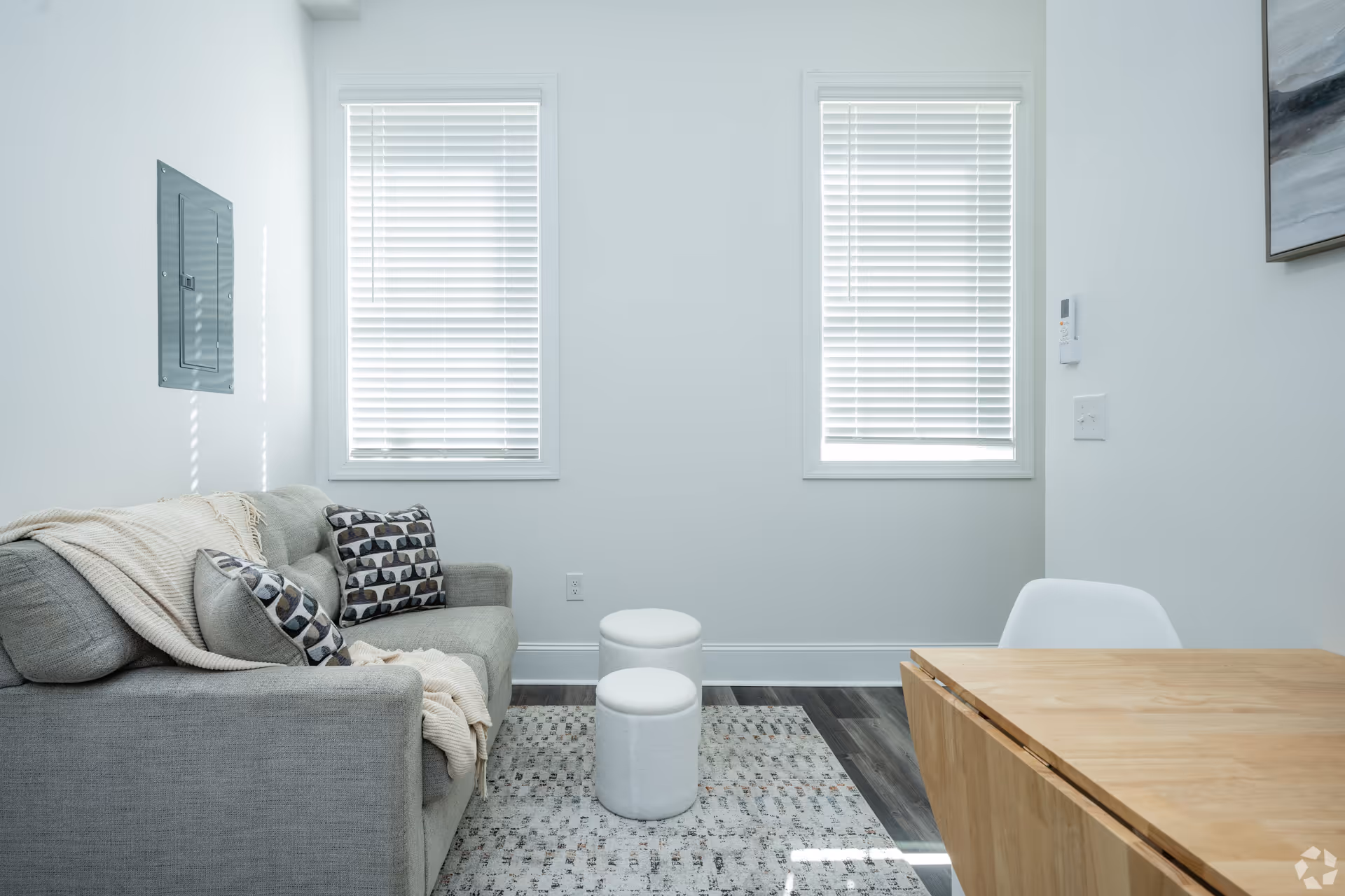 A small living room area with a gray sofa adorned with patterned pillows and a beige throw blanket. Two white round ottomans are placed on a patterned rug in front of the sofa. The room has two windows with white blinds, light-colored walls, and a wooden table with a white chair on the right side. A piece of abstract artwork is partially visible on the right wall.
