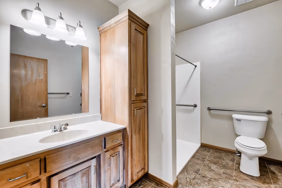 A clean bathroom with a wooden vanity featuring a white countertop and sink, a large mirror above the sink with three light fixtures, a tall wooden cabinet, a walk-in shower with a grab bar, and a toilet with a grab bar on the wall beside it. The floor is tiled in a brown pattern.