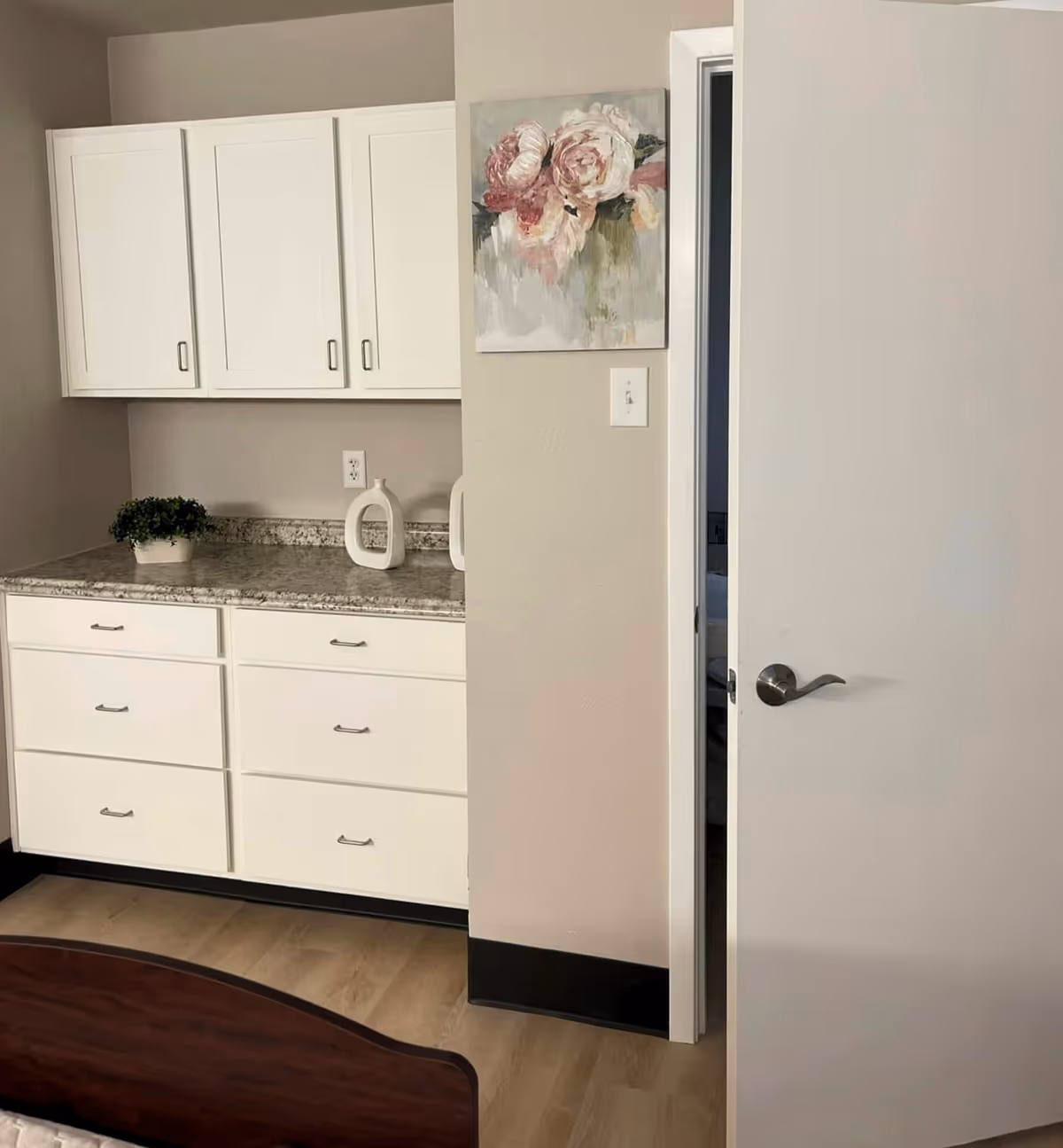 A bedroom corner with white cabinets and drawers, a granite countertop with decorative items, floral wall art, and a partially open door.