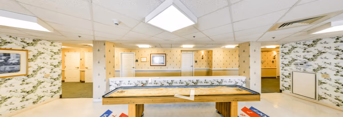 Interior view of a recreational room in an assisted living facility featuring a shuffleboard table in the center. The walls are decorated with patterned wallpaper and framed pictures. There are multiple doors leading to other rooms, and the ceiling has fluorescent lighting panels.