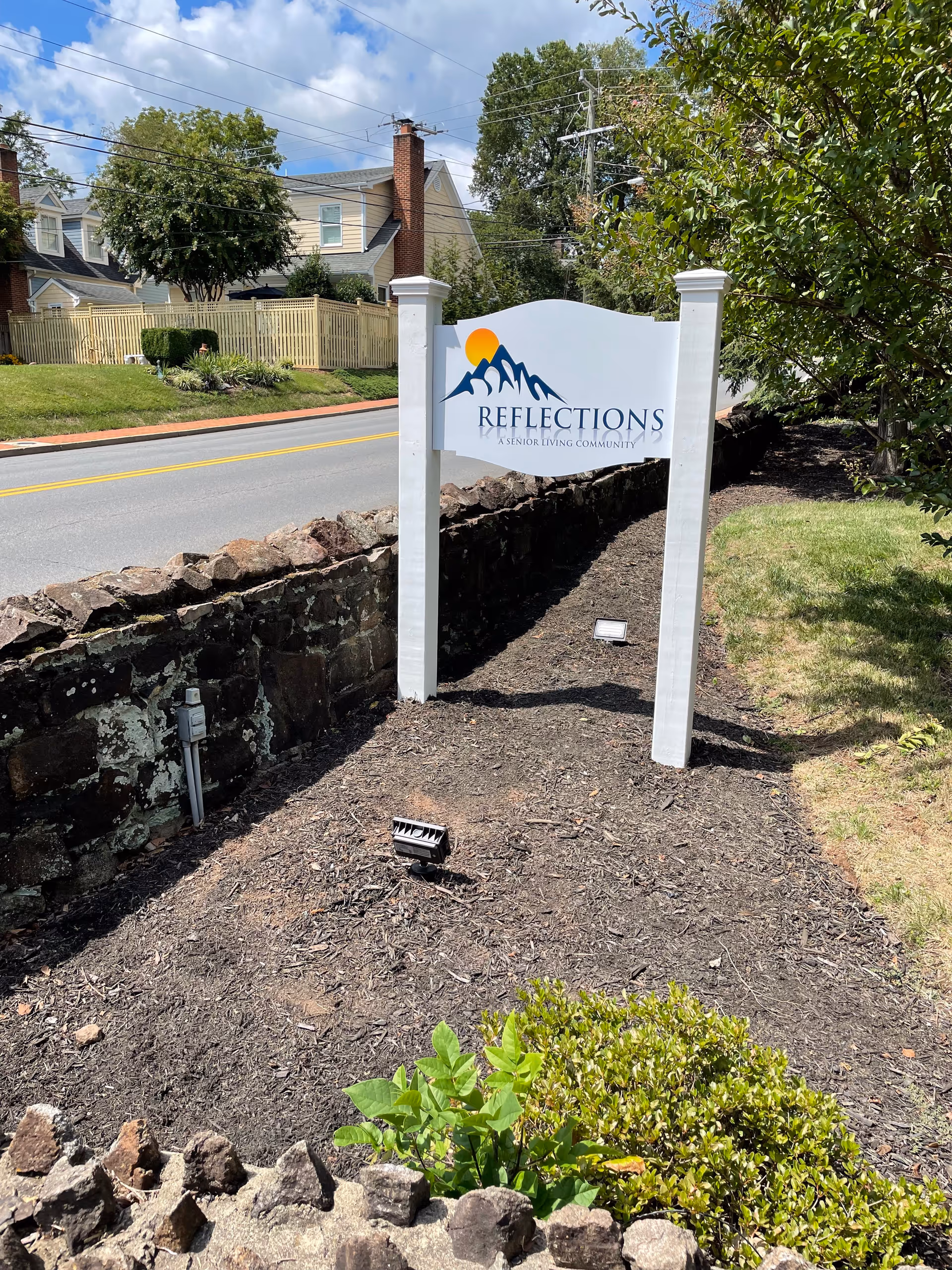 Outdoor view of a white sign with the logo and text 'Reflections A Senior Living Community' mounted on two white posts, situated next to a stone wall along a road with houses and trees in the background under a partly cloudy sky.