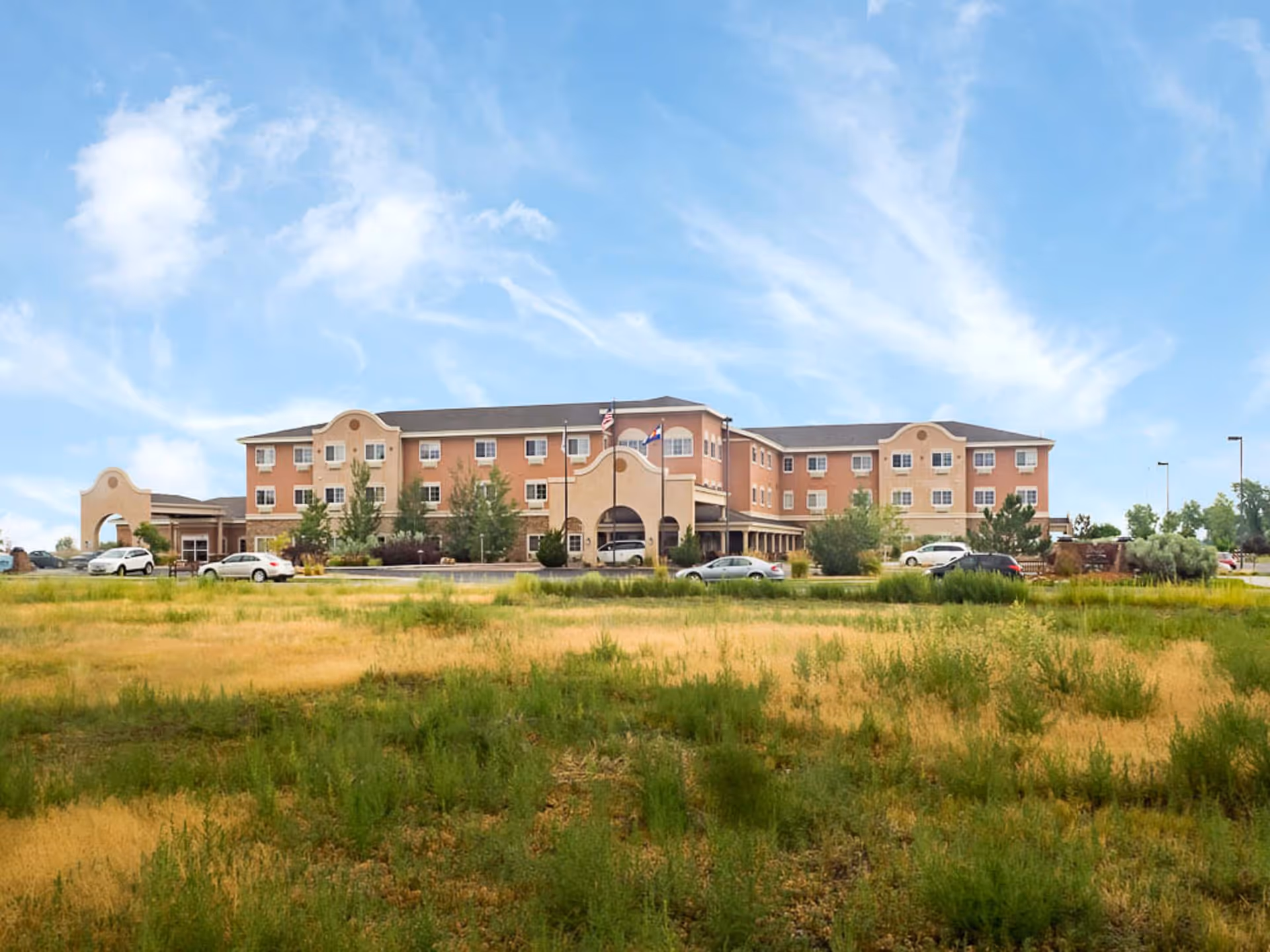 Exterior view of The Bridge at Alamosa, a three-story senior living facility with a beige and light brown facade, multiple windows, and two flagpoles in front. Several cars are parked near the entrance, and there is a grassy field in the foreground under a partly cloudy blue sky.
