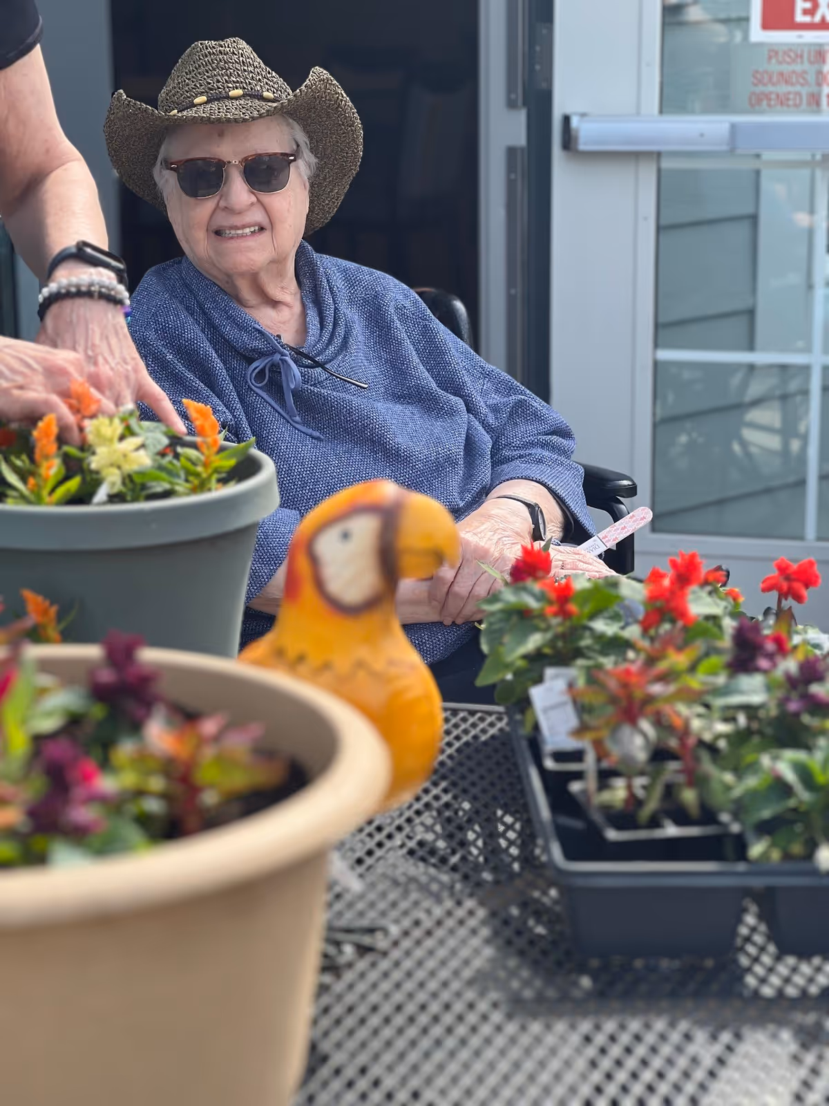 An elderly woman wearing a straw hat and sunglasses is sitting in a wheelchair outdoors, smiling. She is surrounded by potted flowers and plants on a table, with another person’s hands visible tending to the plants.