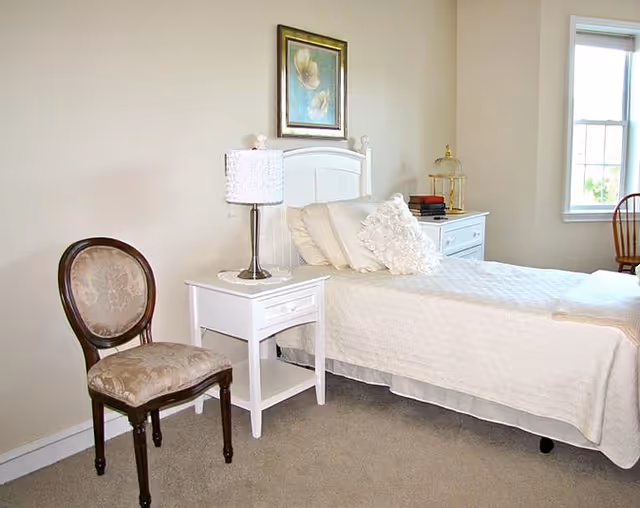A bedroom with a single bed covered in a white quilt and multiple pillows. Next to the bed is a white nightstand with a lamp and decorative items. A wooden chair with a cushioned seat is placed beside the nightstand. There is a framed floral artwork hanging on the beige wall above the bed. A window with white trim is visible on the right side, letting in natural light.