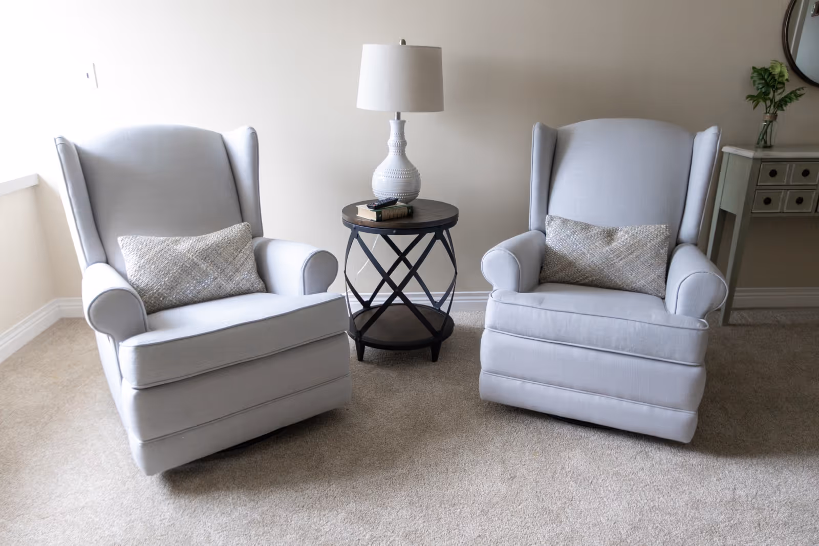 Two light gray upholstered armchairs with matching decorative pillows are positioned on a beige carpeted floor with a round wooden side table between them. The side table holds a white table lamp and a couple of books. In the background, there is a small console table with drawers and a vase with green foliage on top against a plain light-colored wall.